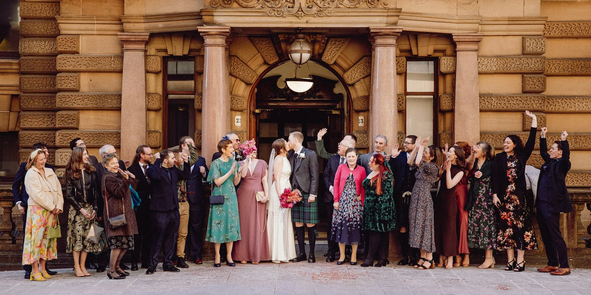 A group of people gathered outside a brownstone with ornate architectural details for a wedding celebration, featuring a bride and groom in the center, surrounded by friends and family celebrating and taking photos.