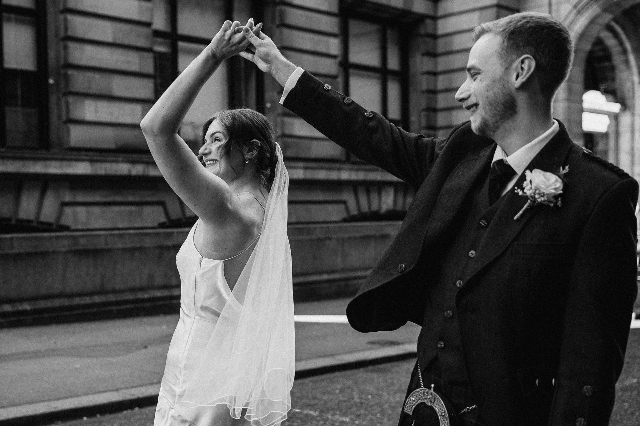 Black and white photo of a happy bride and groom dancing outside on a city street, smiling and holding hands.