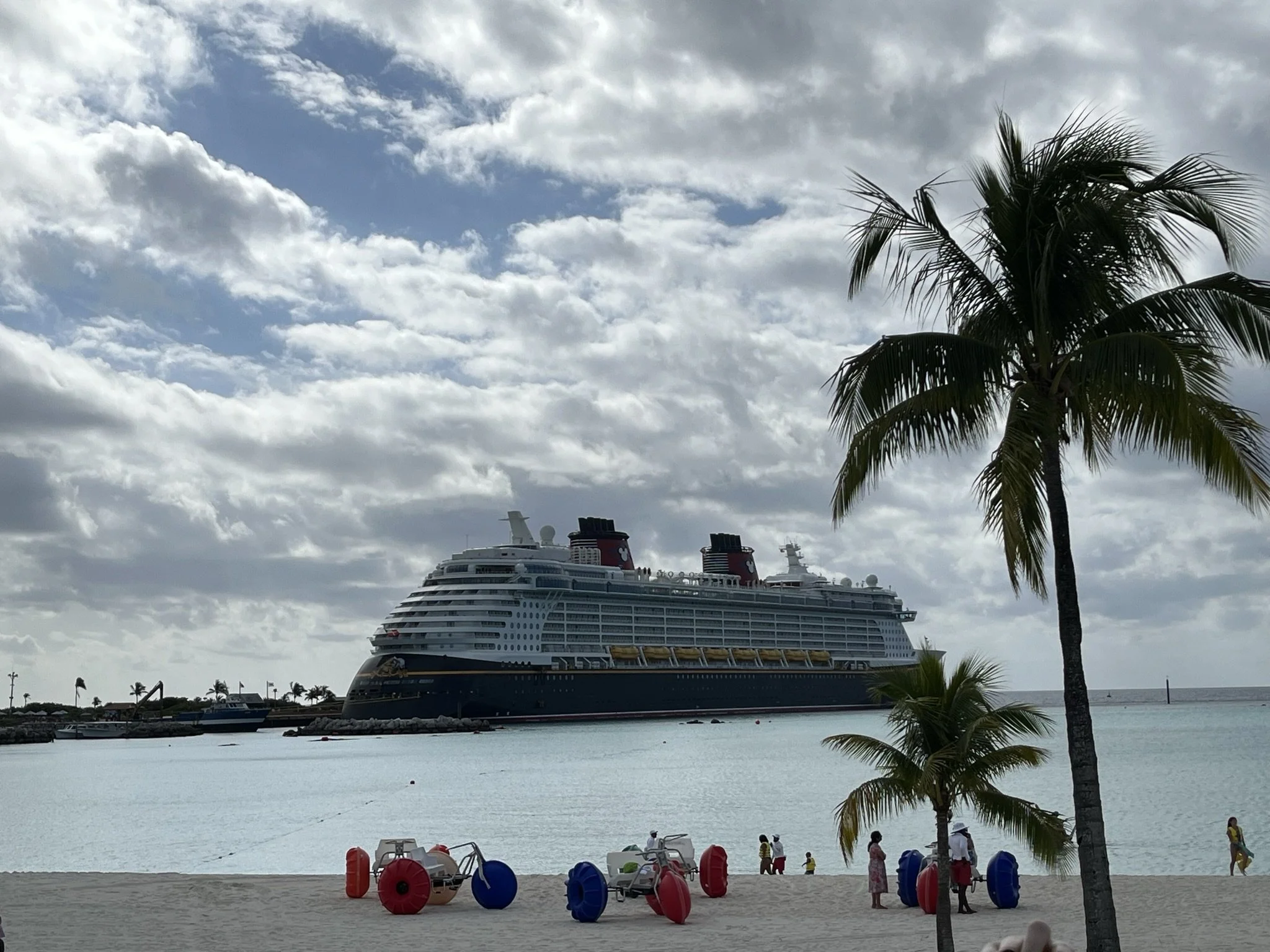 Een groot cruiseschip ligt voor de kust in een tropische locatie, met palm bomen op het strand en enkele mensen die langs de waterkant lopen. De lucht is gedeeltelijk bewolkt.