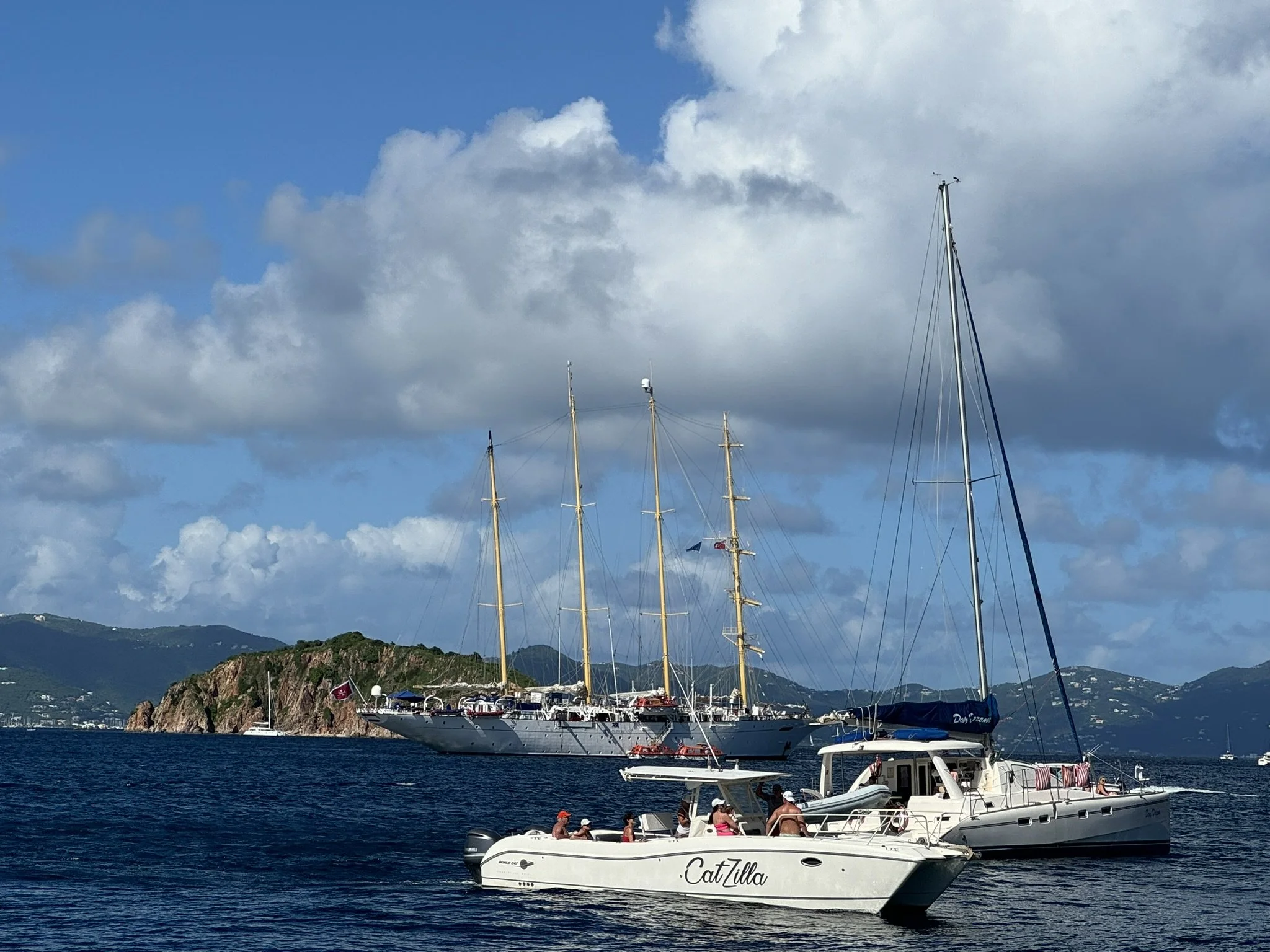 Drie jachten op het water met een bergachtig landschap op de achtergrond en een bewolkte blauwe lucht.