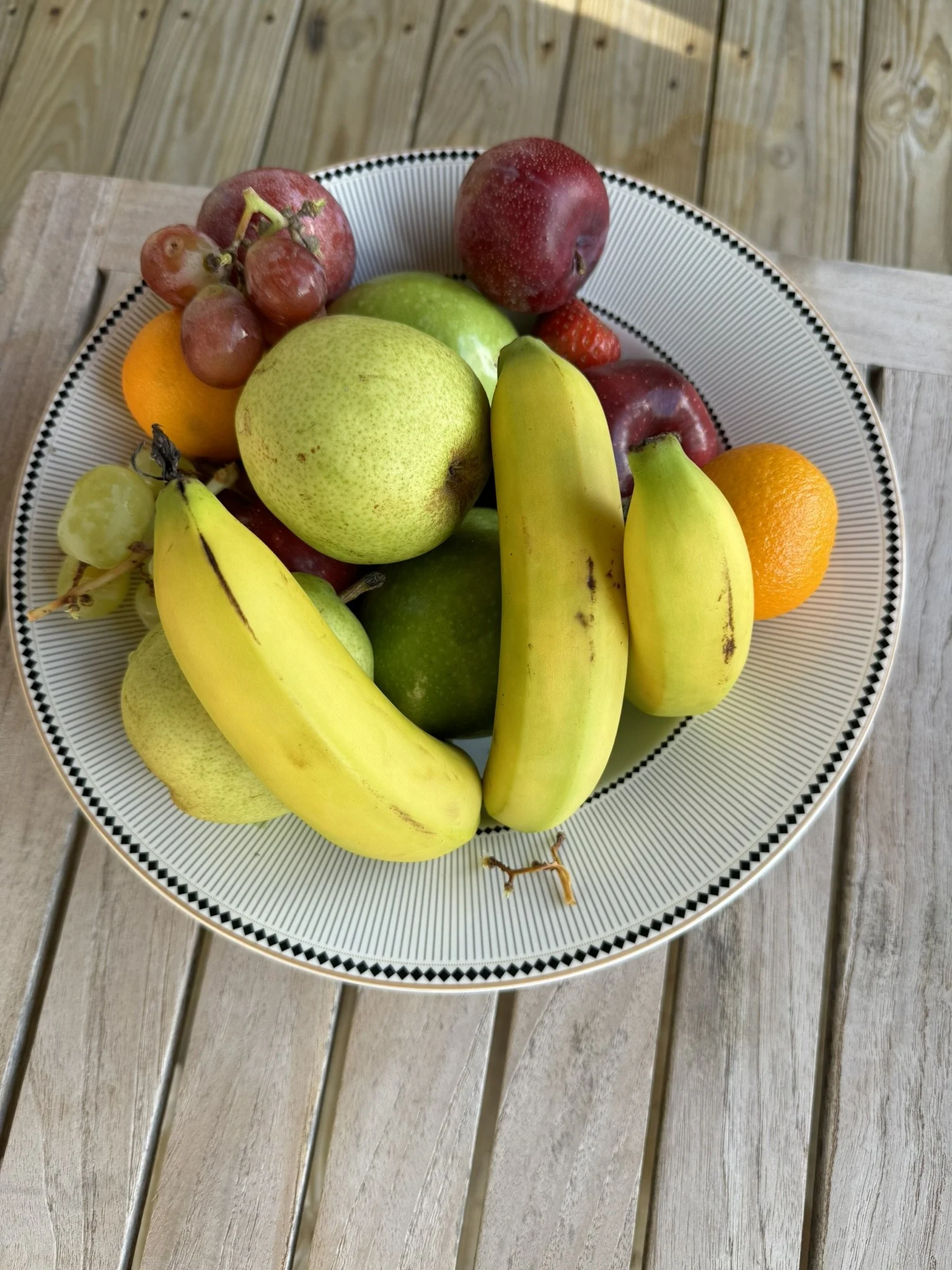 Een witte schaal met een rand met een zwart-wit patroon, gevuld met verschillende soorten fruit zoals bananen, groene appels, rode appels, druiven, een sinaasappel, en aardbeien,op een houten tafel.