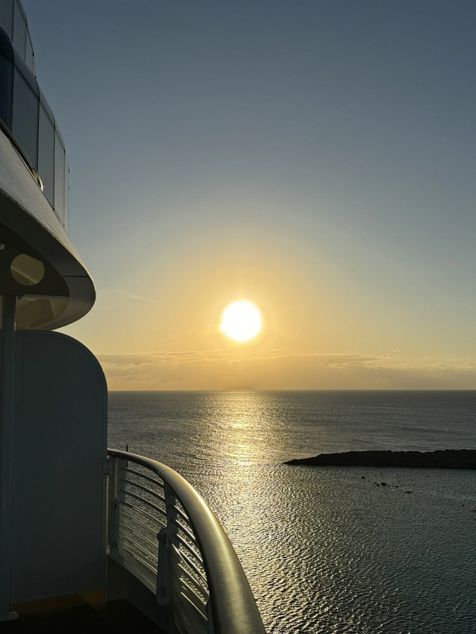 De foto toont een zonsondergang boven de oceaan, genomen vanaf het balkon van een cruiseschip. Aan de linkerkant zijn delen van het glazen raam en de reling van het schip zichtbaar.