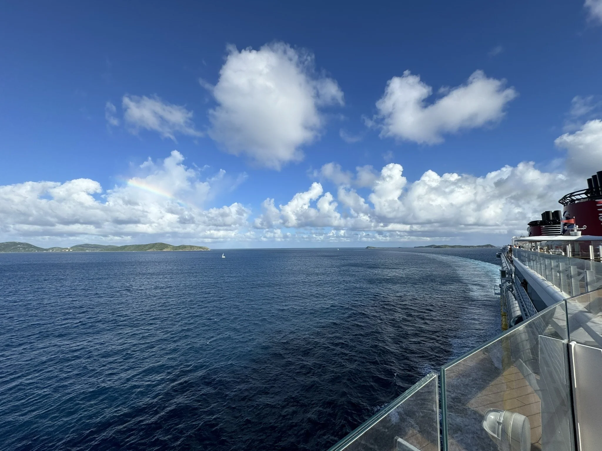 Uitzicht vanaf een schip op de oceaan, met eilandjes op de achtergrond en een regenboog in de lucht, de scheepsromp en reling zijn zichtbaar aan de rechterkant.
