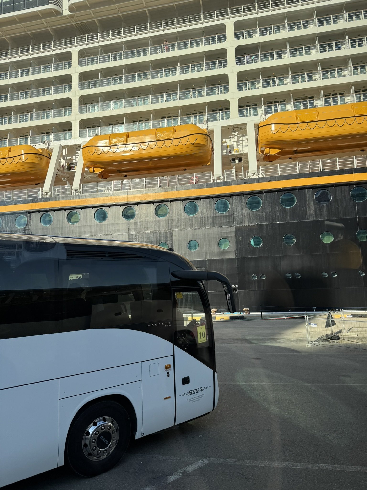 Een wit busje geparkeerd op een parkeerplaats met een cruiseschip op de achtergrond, dat meerdere verdiepingen en gele lifeboons heeft.