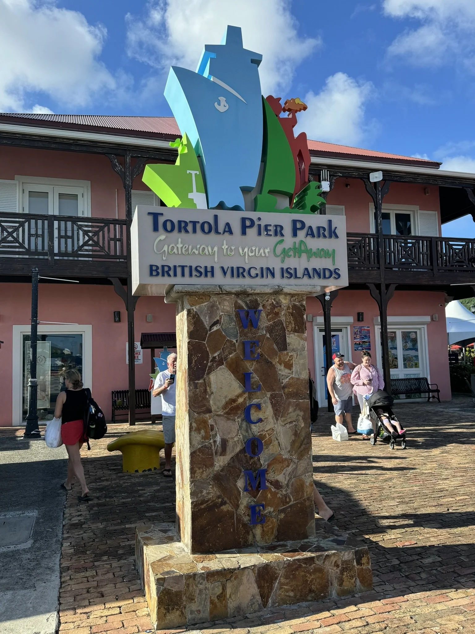 Ingangsbord met naam Tortola Pier Park en een tekst dat de toegangspoort naar de Britse Maagdeneilanden is, met kleurrijke grafische weergaven van een schip, een zee en een vulkaan. Het bord staat op een stenen zuil, met een roze gebouw en mensen erb
