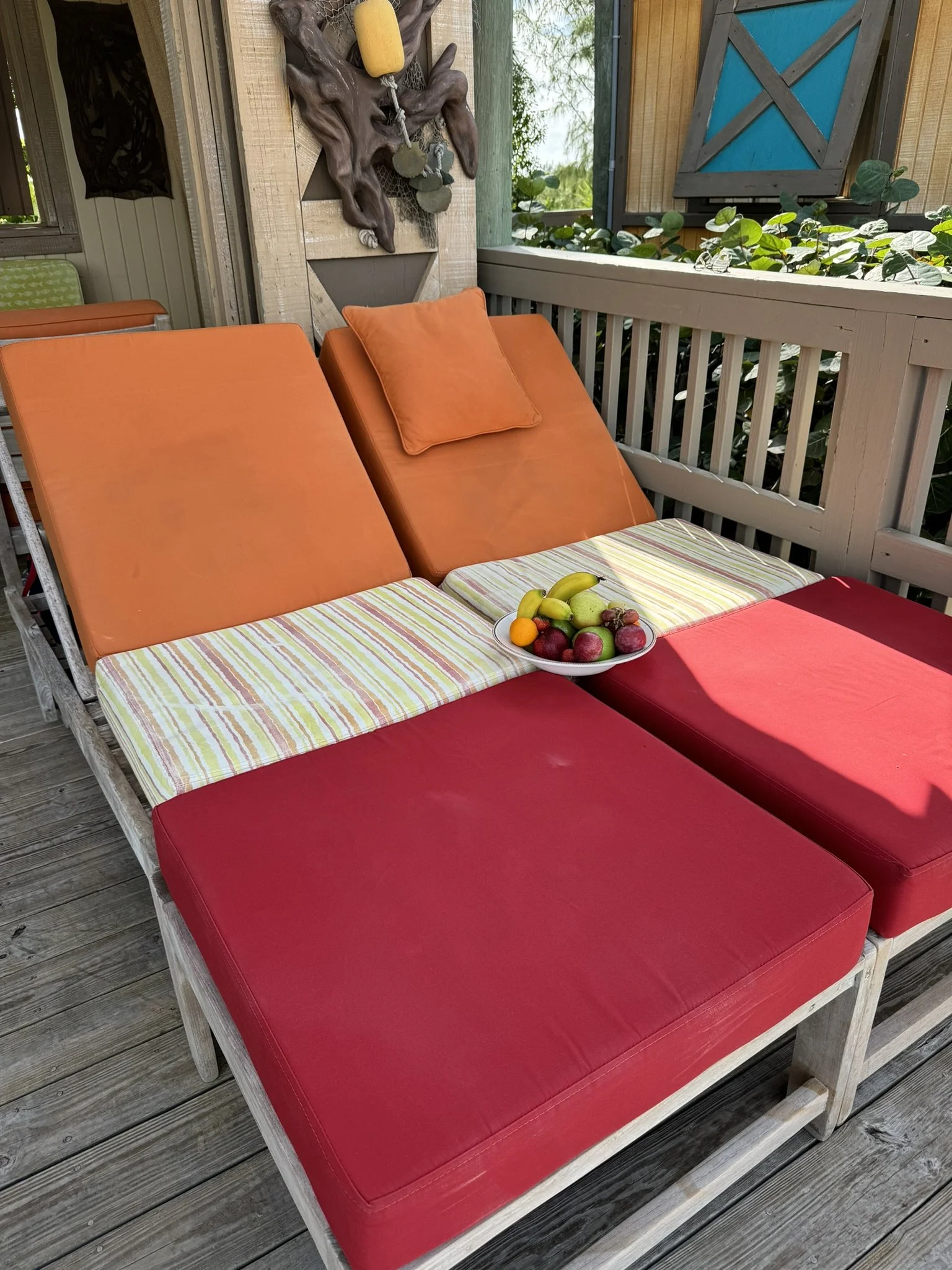 Twee ligbedden met rode en oranje kussens en een bord met fruit op een houten veranda, met een houten hek en planten erachter.