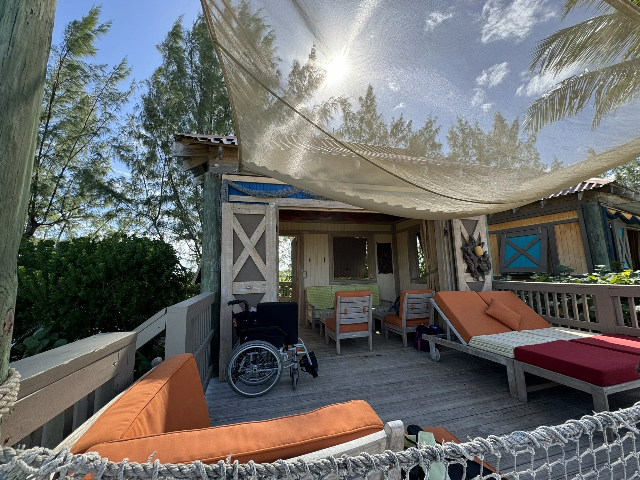 Buiten terras met schommelstoel, fauteuils en een bed met oranje kussens, onder een witte zonnescherm, omgeven door groene bomen.