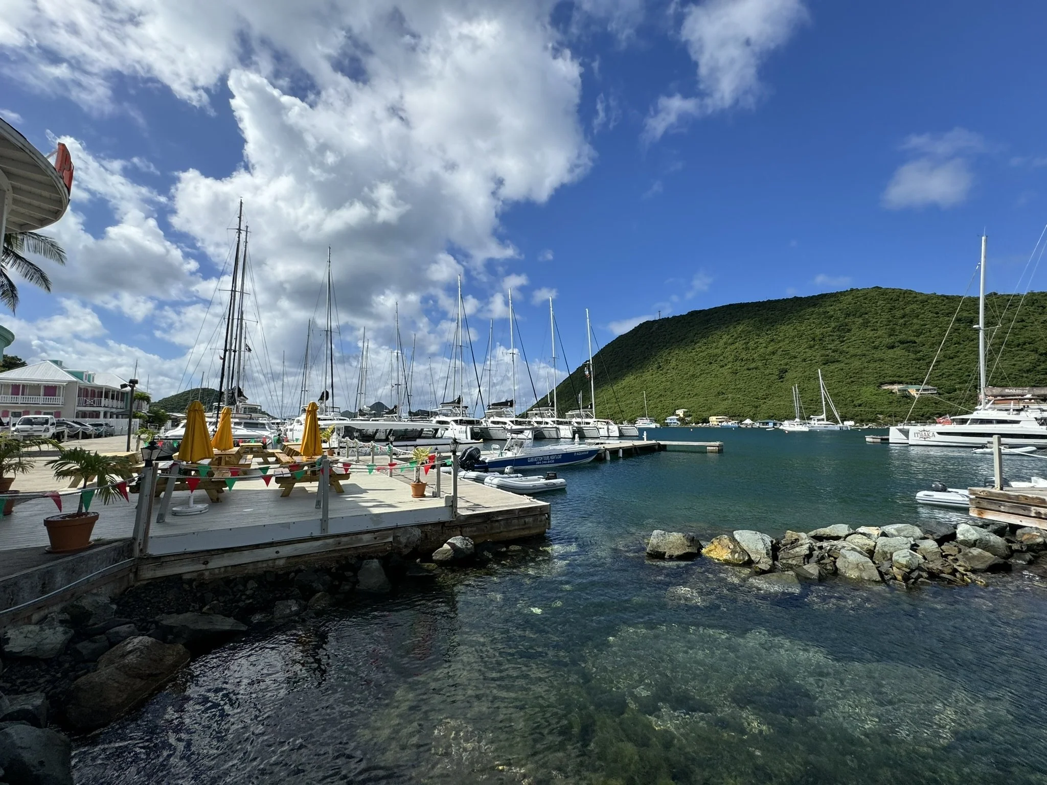 Een haven met boten en sailboats, groene heuvels op de achtergrond, blauwe lucht met wolken, terras met gele parasols.