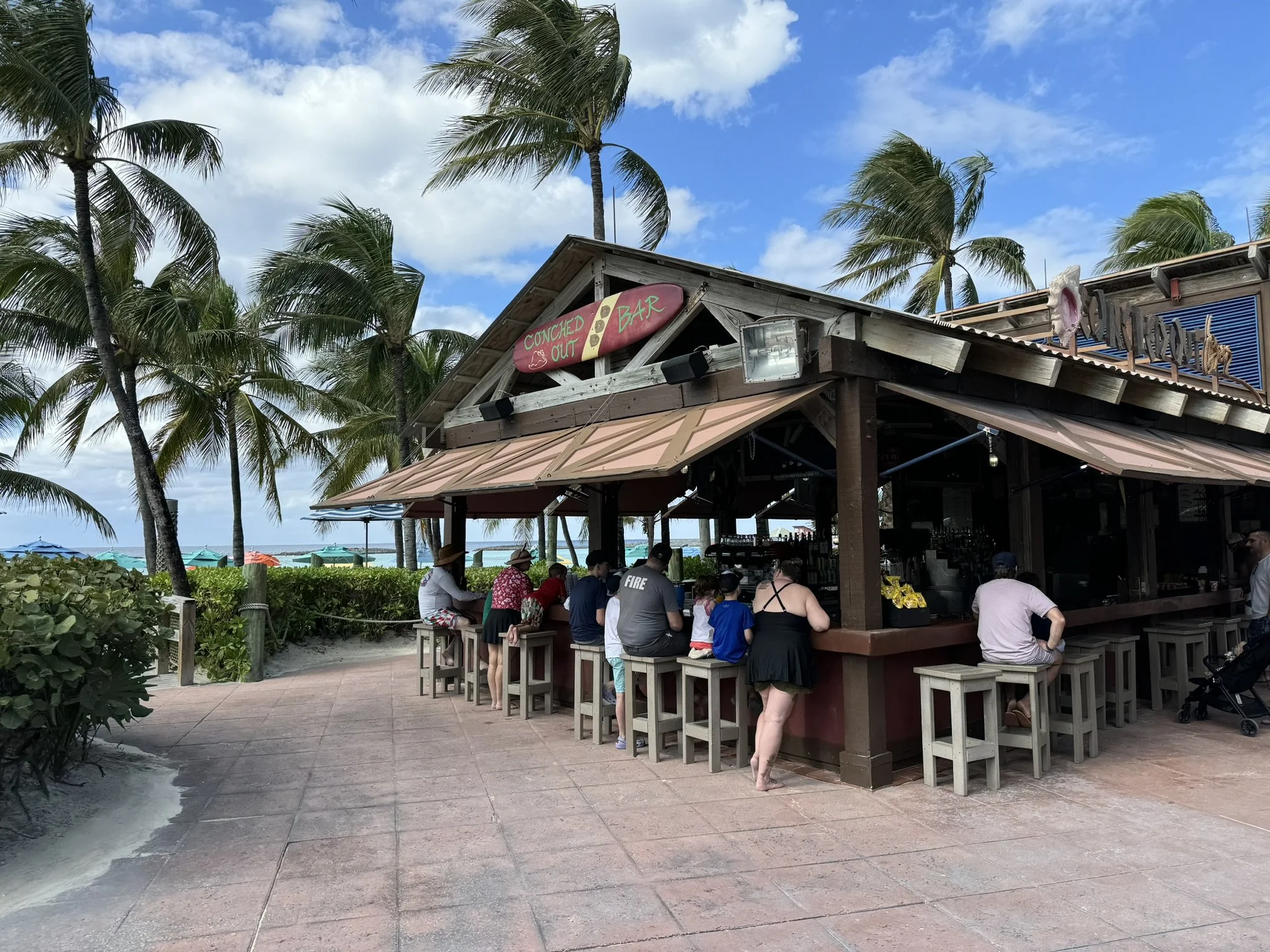 Bar op het strand met mensen die genieten van hun drankje, omgeven door palm bomen en blauwe lucht.