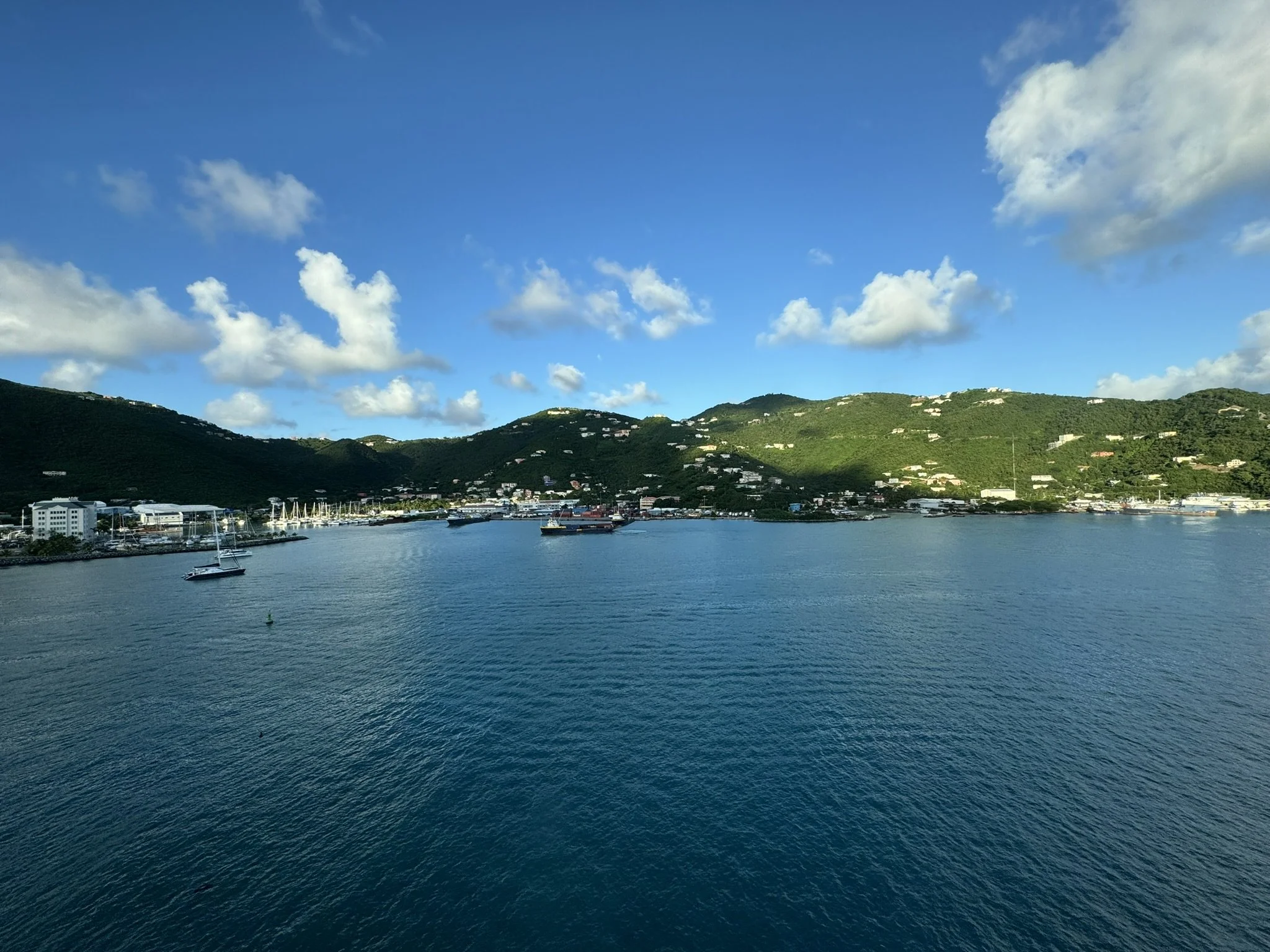 Uitzicht op een waterkant met boten en groene heuvels onder een blauwe lucht met wolken.