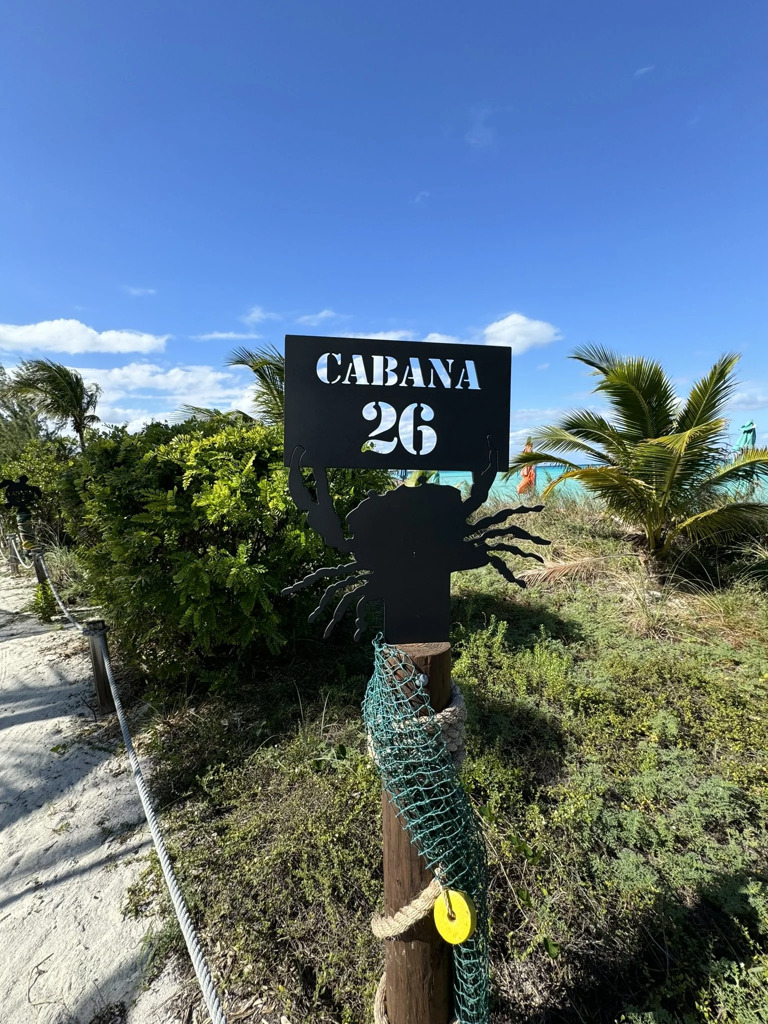Signaalbord met de tekst "CABANA 26" en een silhouet van een zeekreeft, geplaatst in een strandsetting met palmbomen en een helder blauwe lucht.