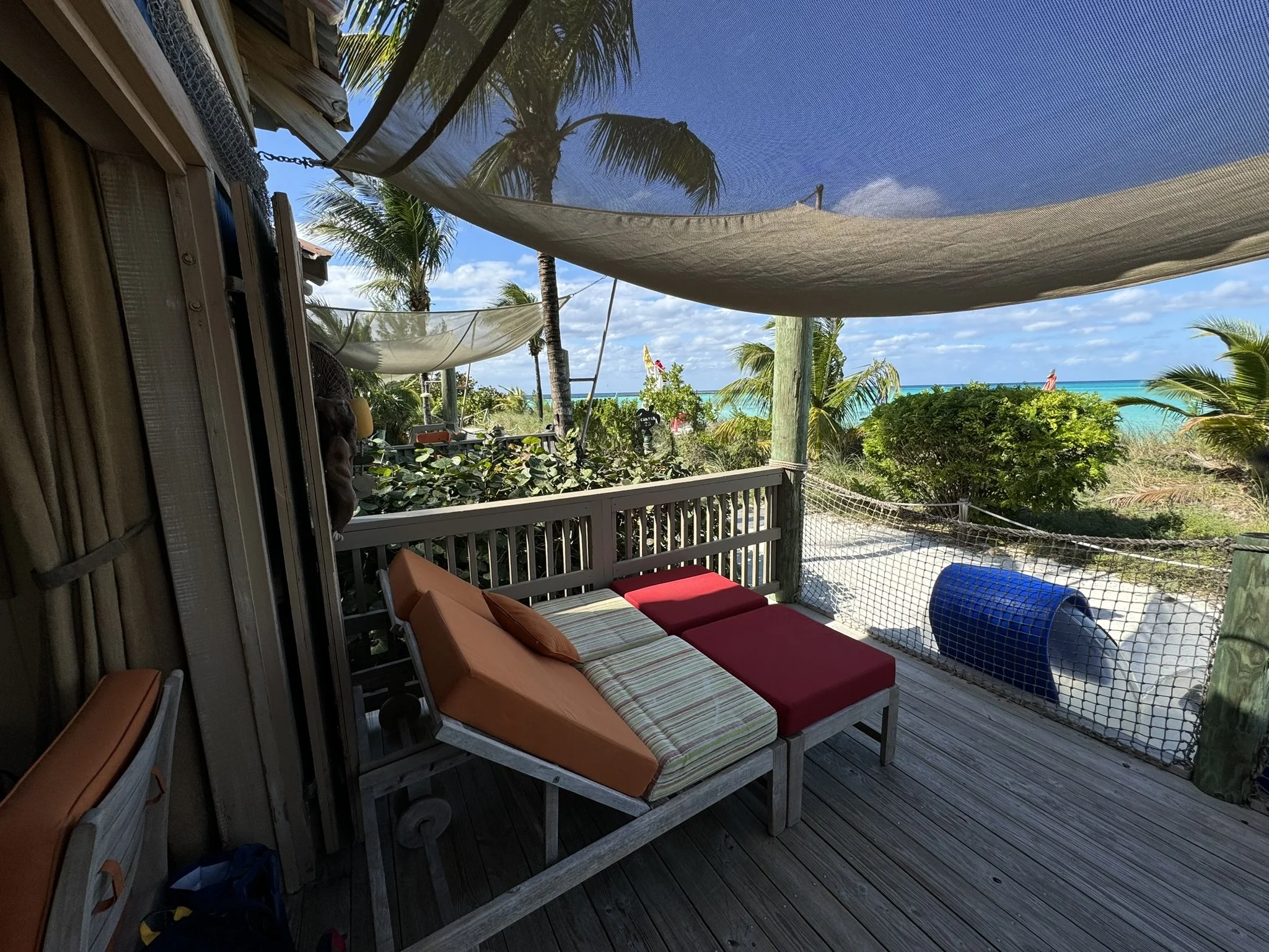 Een zonnig terras met houten vloer en gele en rode ligbedden, omgeven door palmbomen en uitzicht op de oceaan, deels afgeschermd met stof en netten.