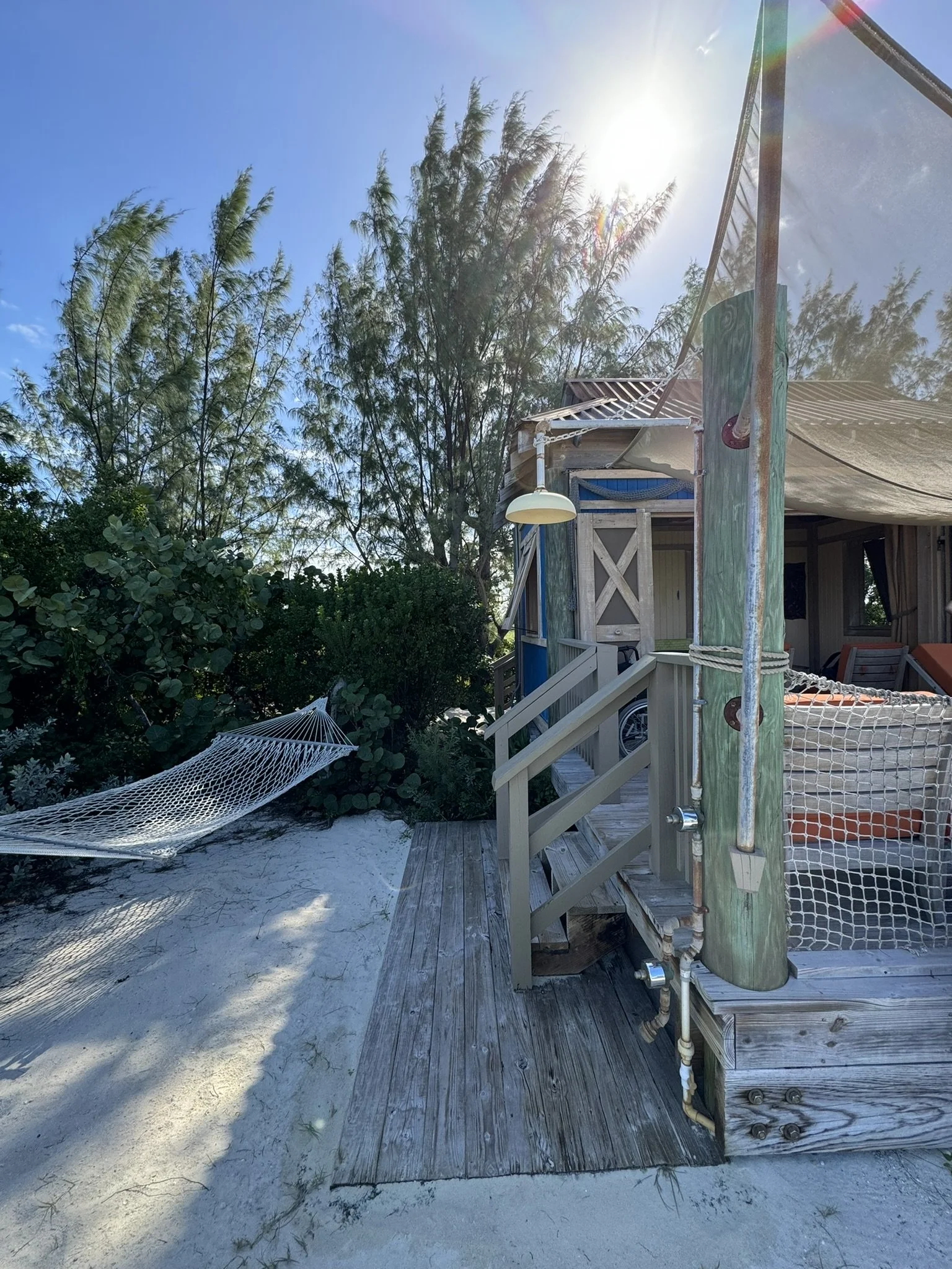 Een houten hut op een strand, met een trap en een hangmat tussen de bomen, onder een heldere blauwe lucht met de zon schijnend.