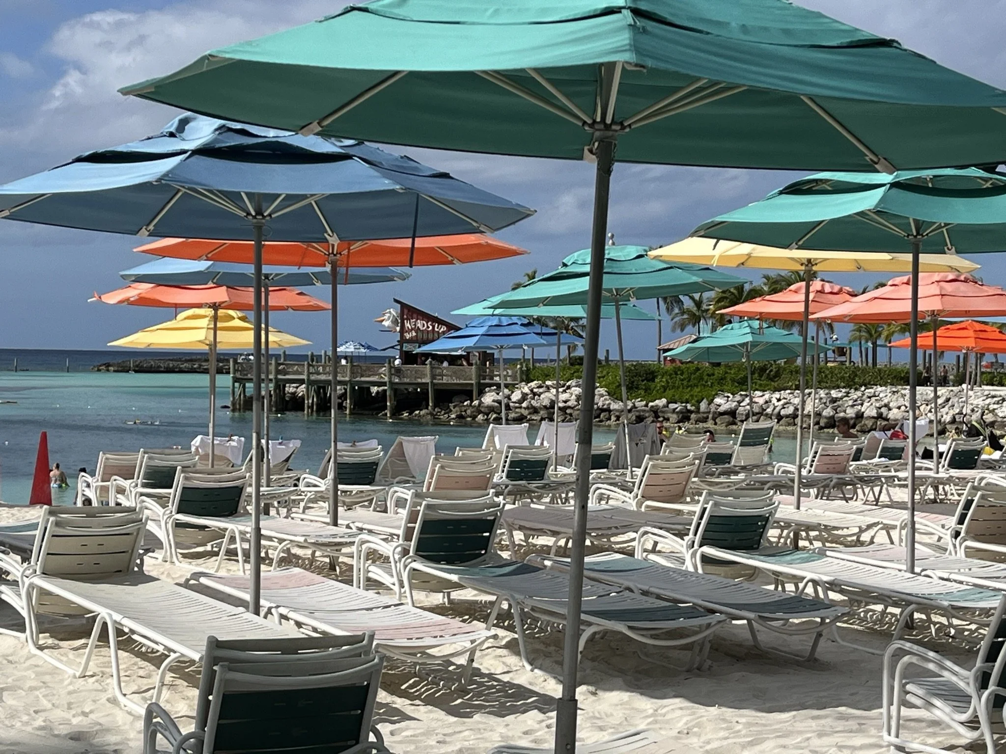 Strand met ligstoelen en kleurrijke parasols, helder water en een pier met boten op de achtergrond, onder een bewolkte hemel.