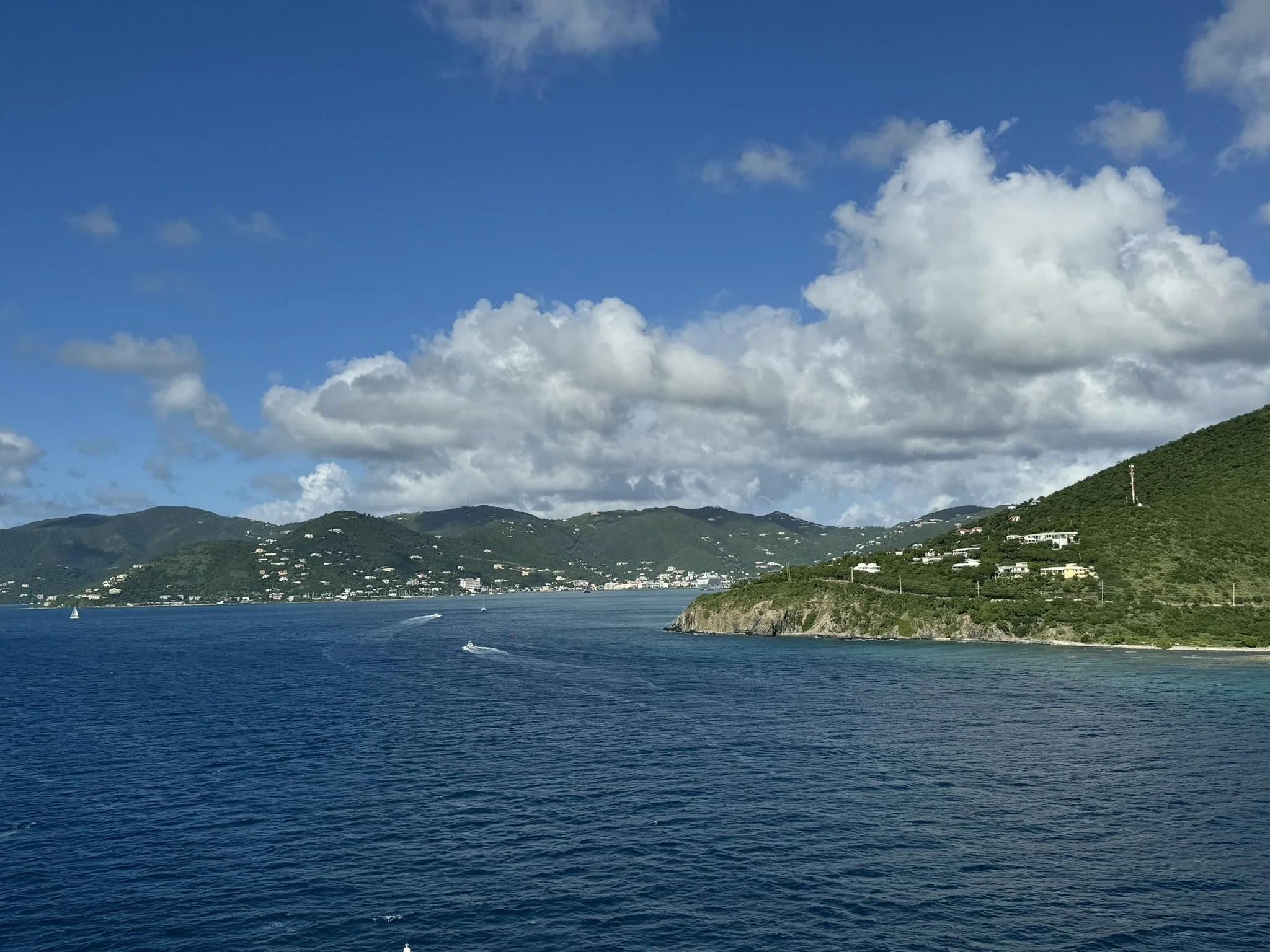 Een foto van een zee met boten en een groen heuvelachtig landschap onder een deels bewolkte blauwe lucht.