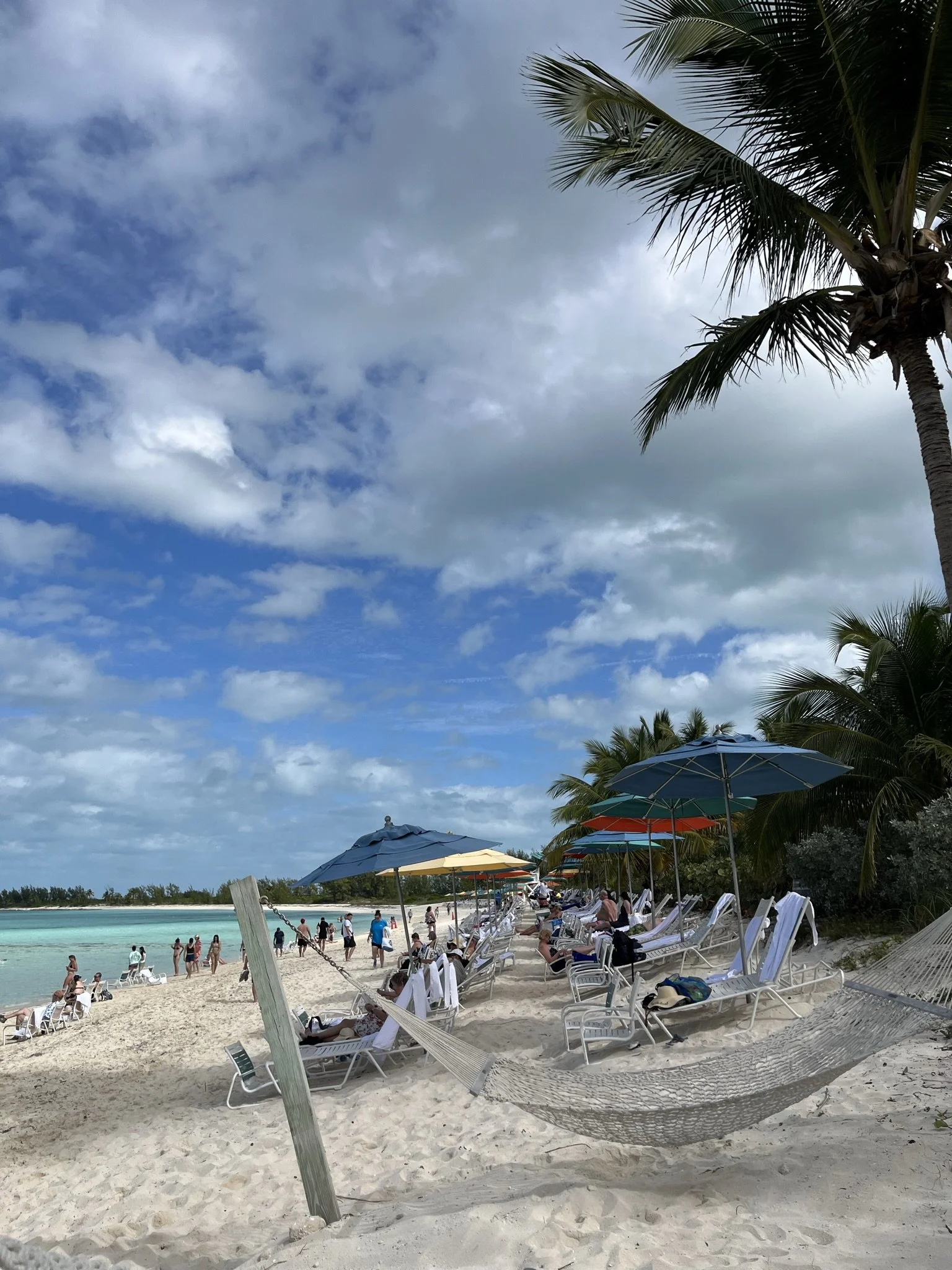 Een strand met ligbedden, parasols, palmbomen en mensen die genieten bij de zee onder een bewolkte lucht.