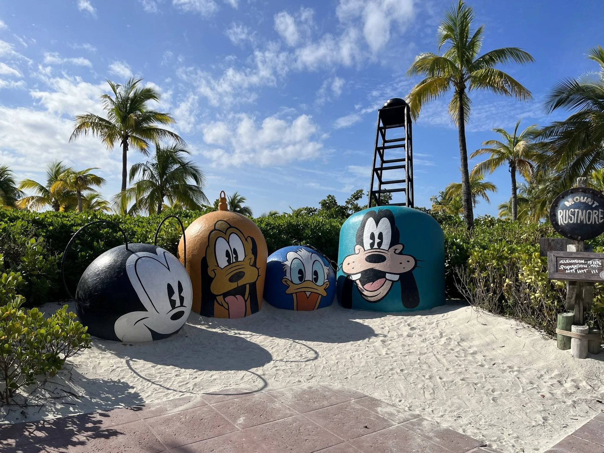 Mickey Mouse, Pluto, Donald Duck en Goofy decoraties op een strand met palm bomen en een blauwe lucht.