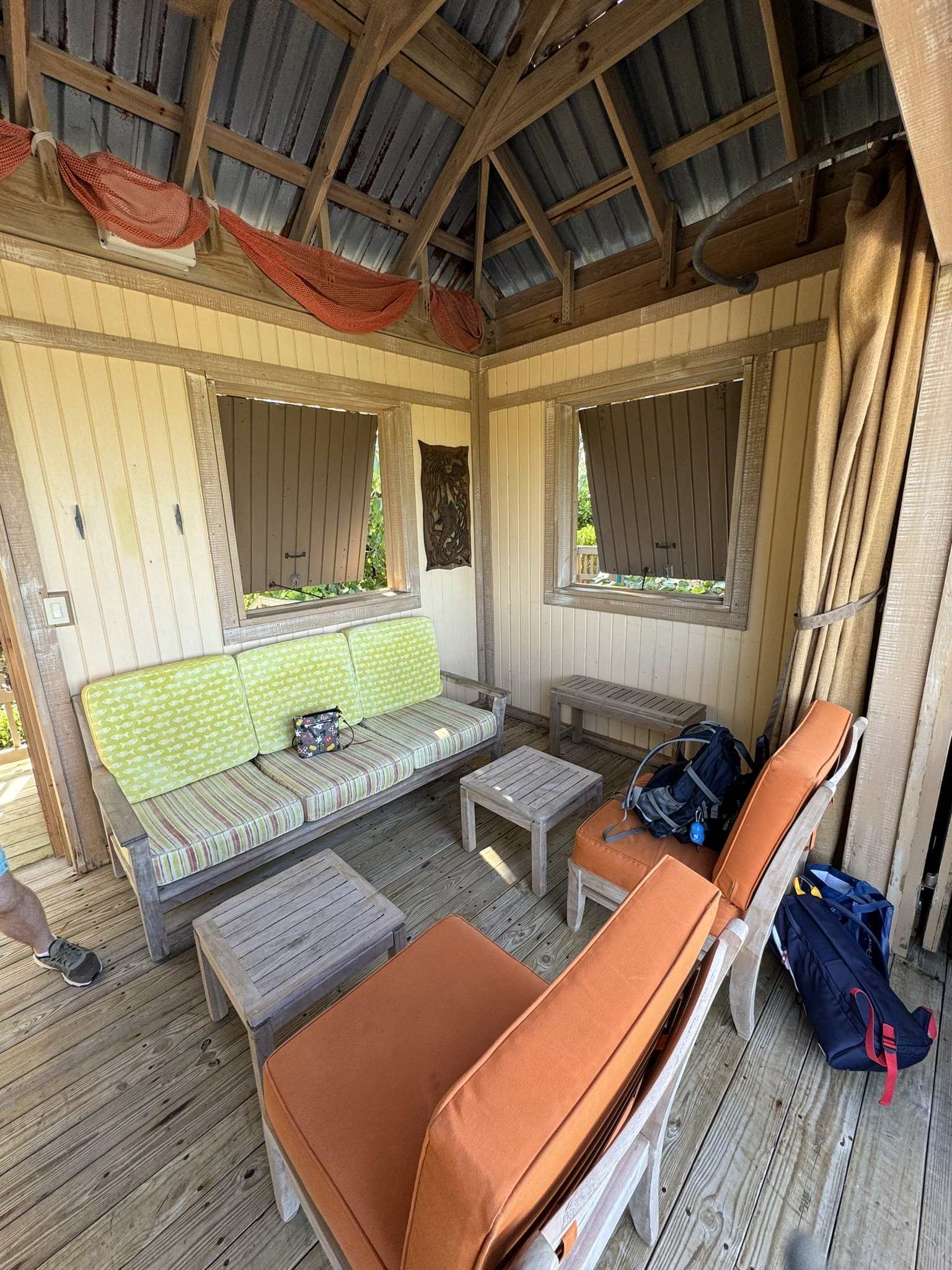 Een rustieke houten veranda met zitplaatsen, waaronder een bank met groene kussens, twee oranje stoelen en bijzettafels. De muren zijn van hout en er zijn twee ramen met luiken, met een open een raam en een gesloten raam. Er hangen decoraties aan de 