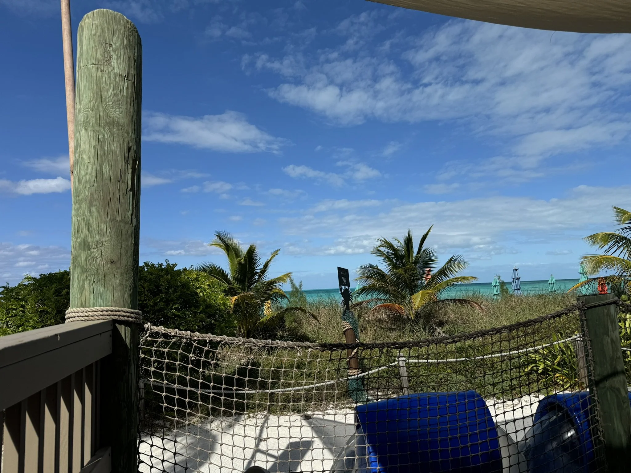 Zicht op een strand met palmbomen, blauwe zee en cabanas op de achtergrond, gezien vanaf een houten terras met een net en blauwe stoelen.