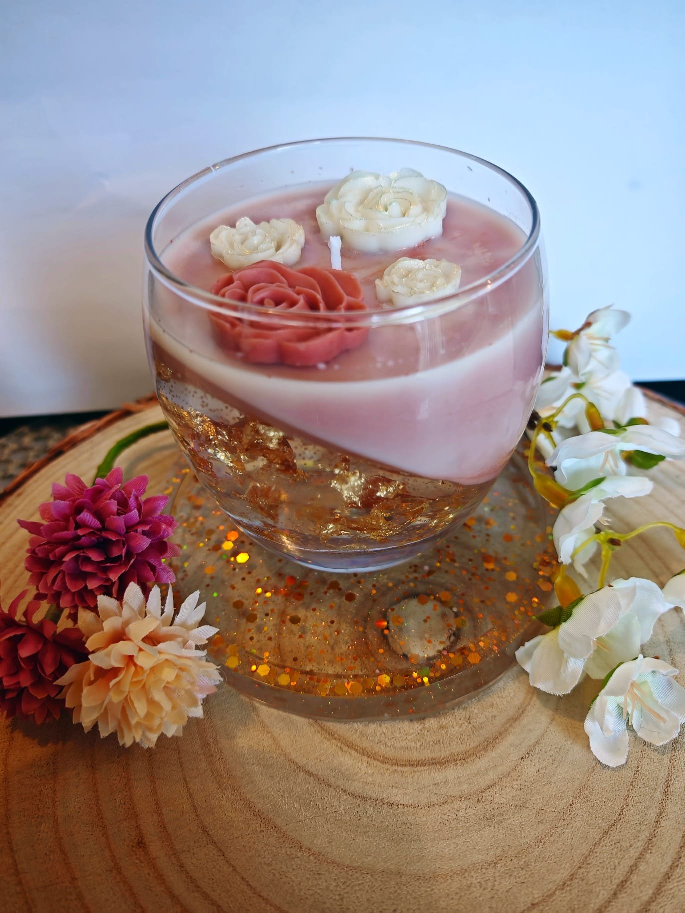 A decorative glass bowl with two layers of liquid, topped with a pink floating candle with white and pink rose-shaped wax decorations. The bowl is on a wooden round board, surrounded by artificial flowers including pink, peach, and white flowers, wit