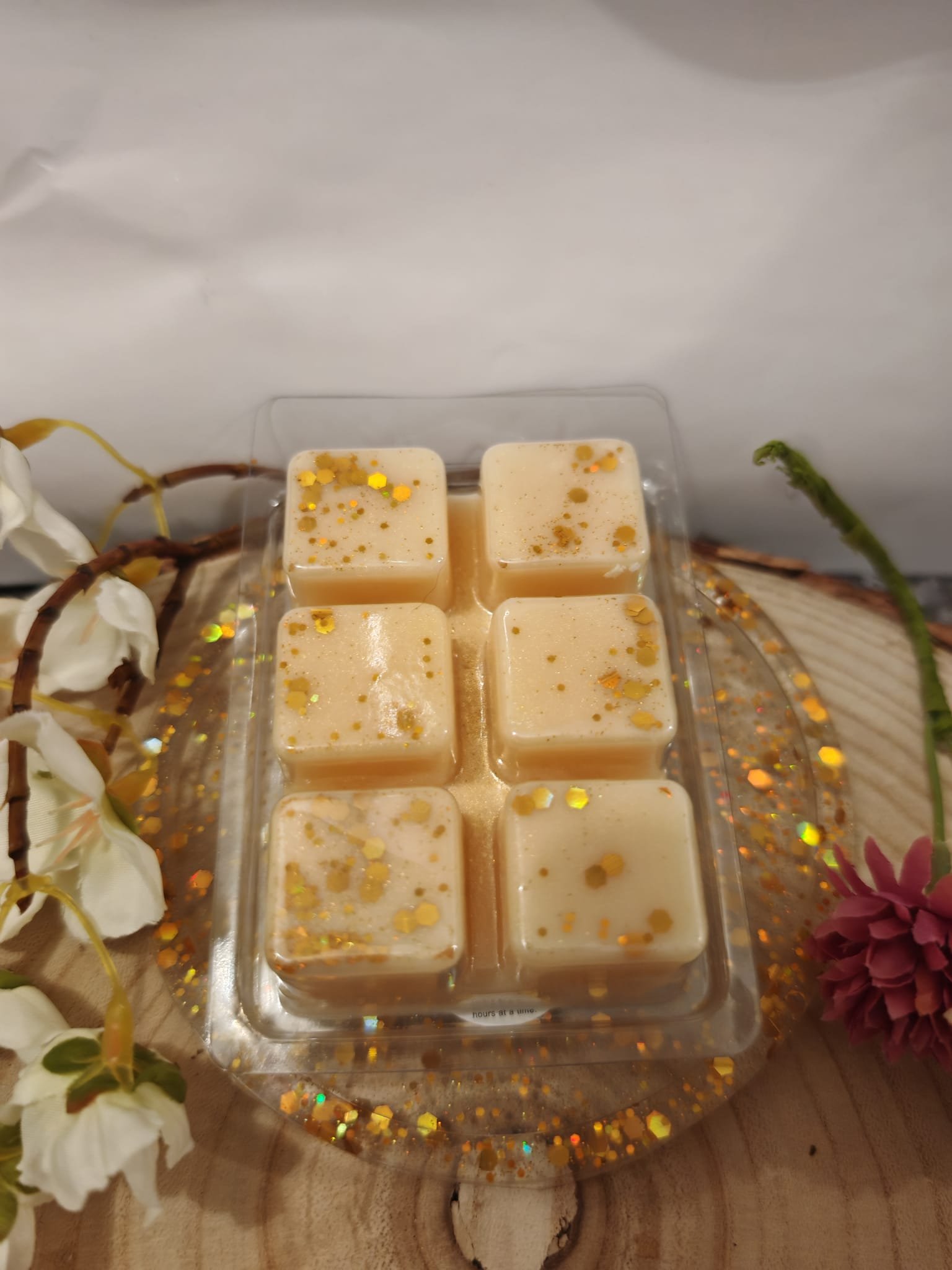 Six creamy white squares of dessert with gold flakes on top, arranged in a plastic container on a wooden surface decorated with white and pink flowers and gold glitter.