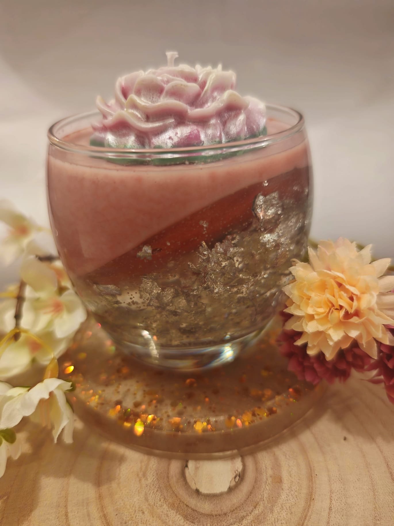 A decorative candle with a pink and white flower inside, placed on a wooden surface with flowers and gold glitter around it.