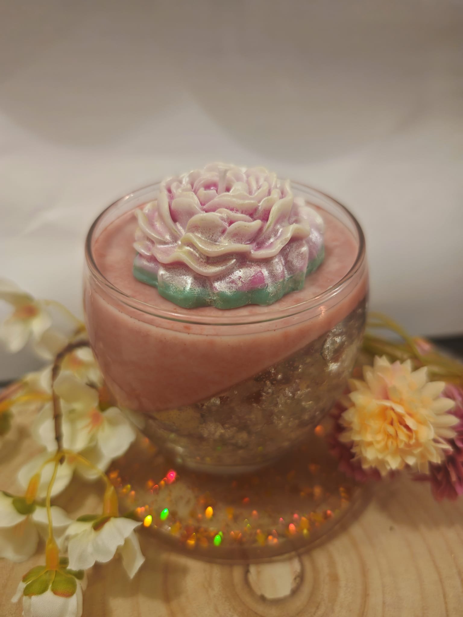 A glass filled with a pink frothy beverage, topped with an intricate 3D flower decoration made of edible material. Surrounding the glass are some colorful flowers and a wooden surface with small, multicolored lights.
