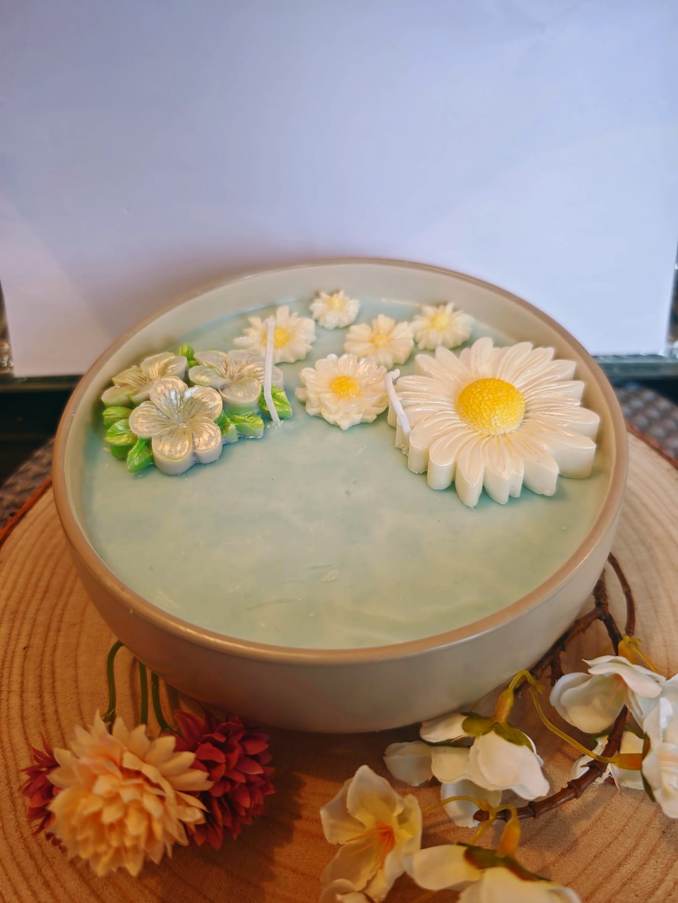 A round white candle with floral designs on top, sitting on a wooden surface with flower decorations around it.