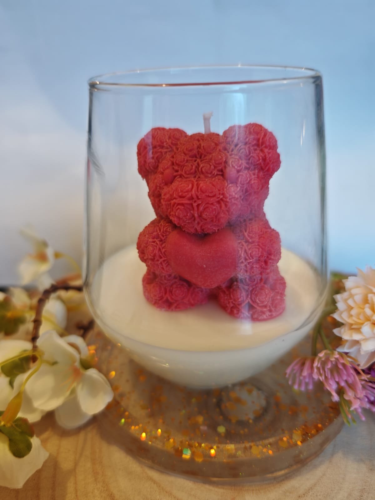 A pink teddy bear-shaped candle inside a glass container, placed on a round, glittery, gold-colored tray with floral decorations around it.