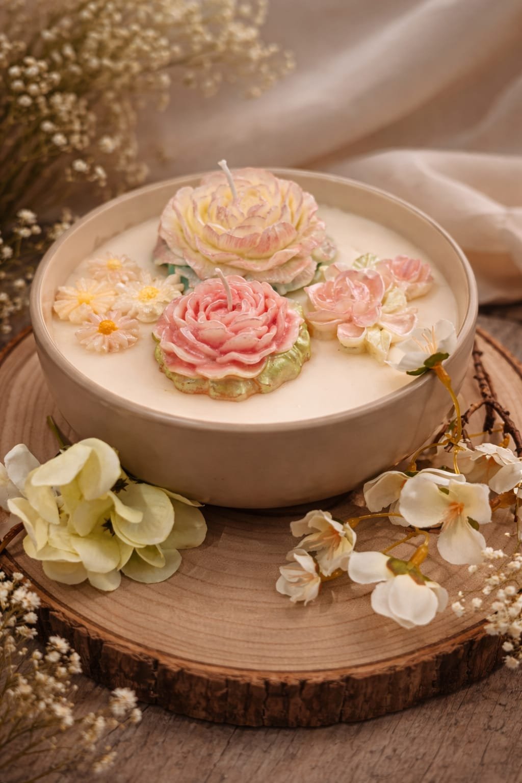 Decorative floral candles on a wooden slice with white and pink flower designs, surrounded by real white and cream flowers.