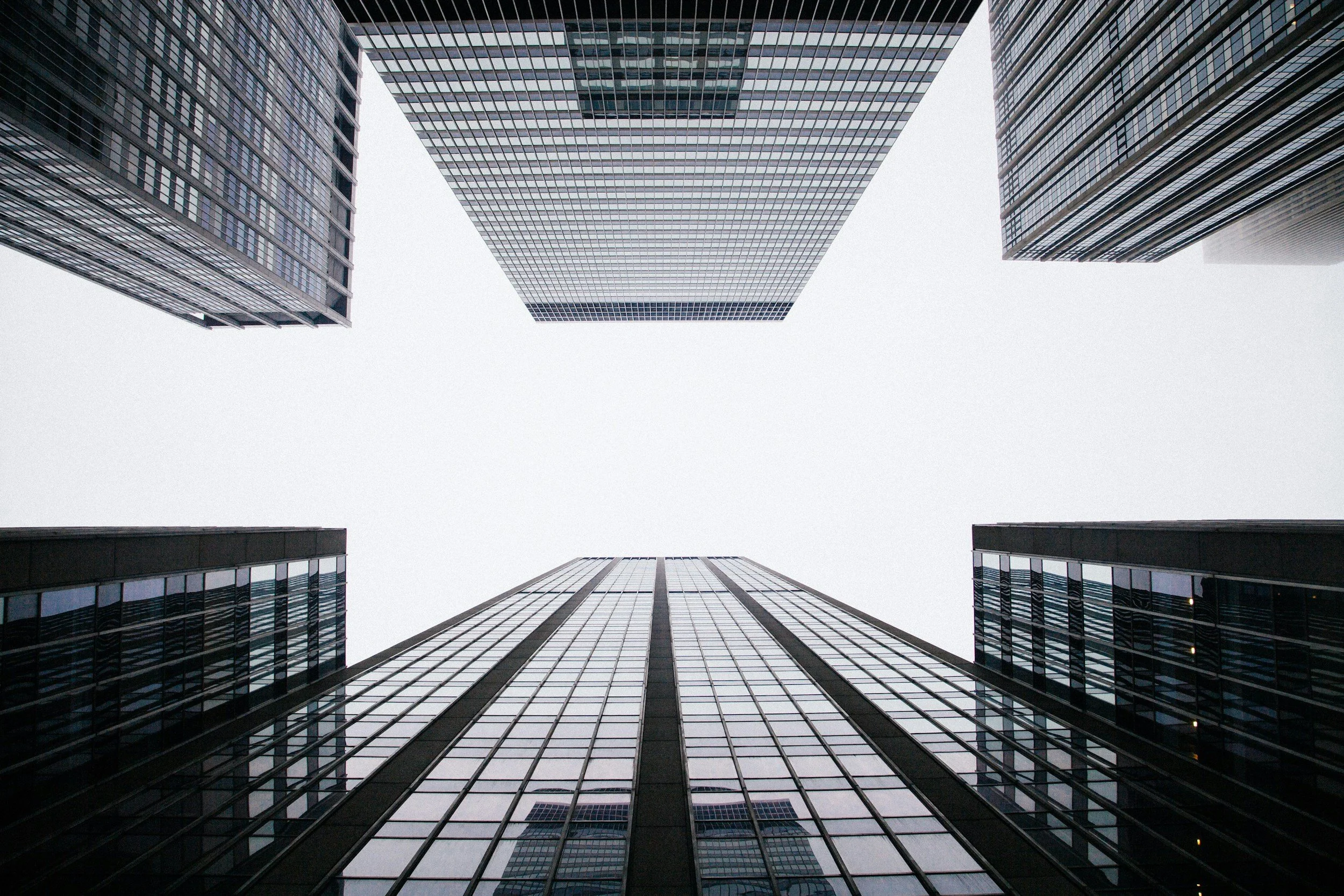 Looking up at skyscrapers in a city, with a view of the overcast sky between modern glass and steel buildings.