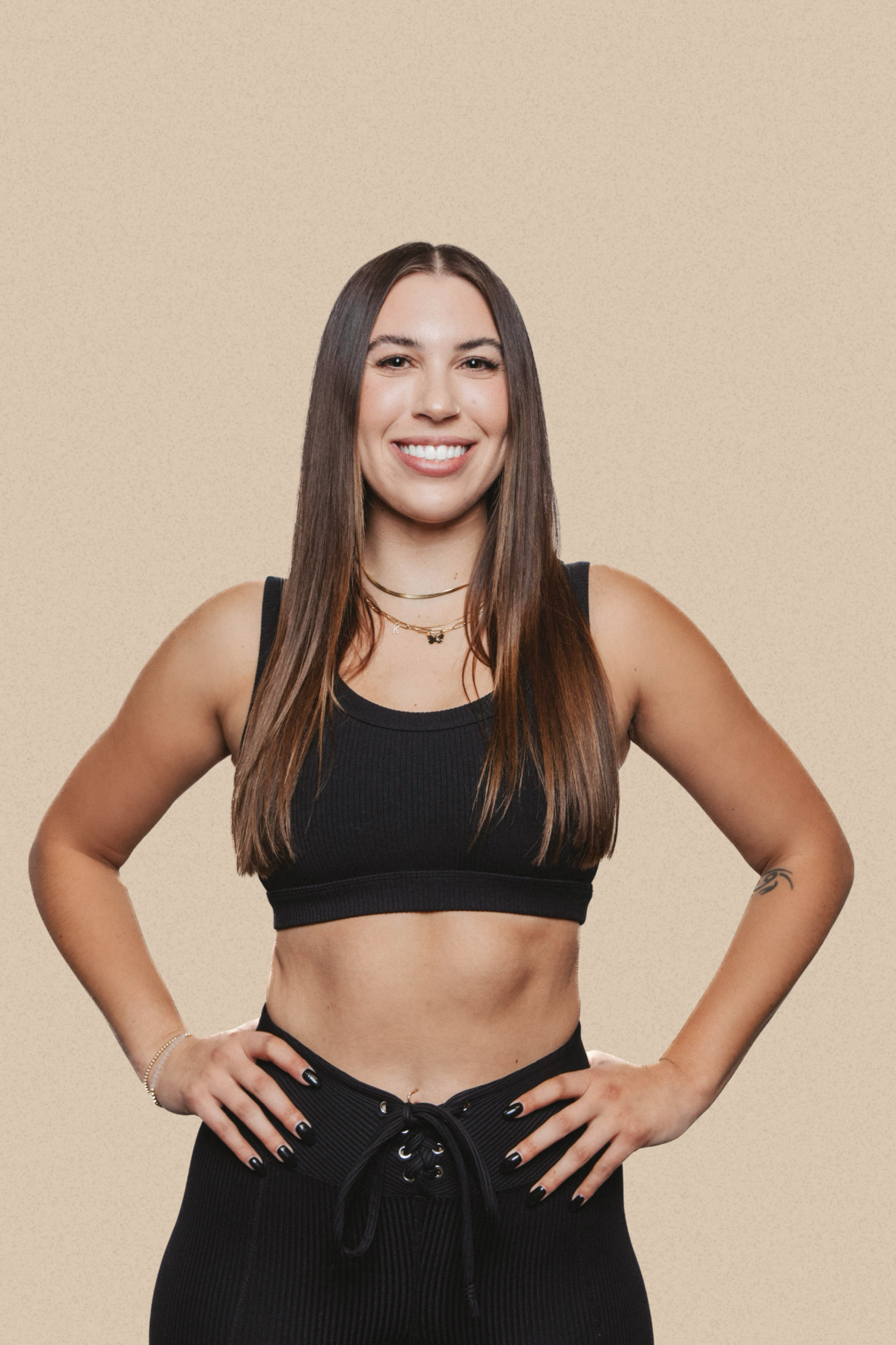 A young woman with long brown hair, wearing a black sports bra and black high-waisted pants, standing with her hands on her hips against a beige background, smiling