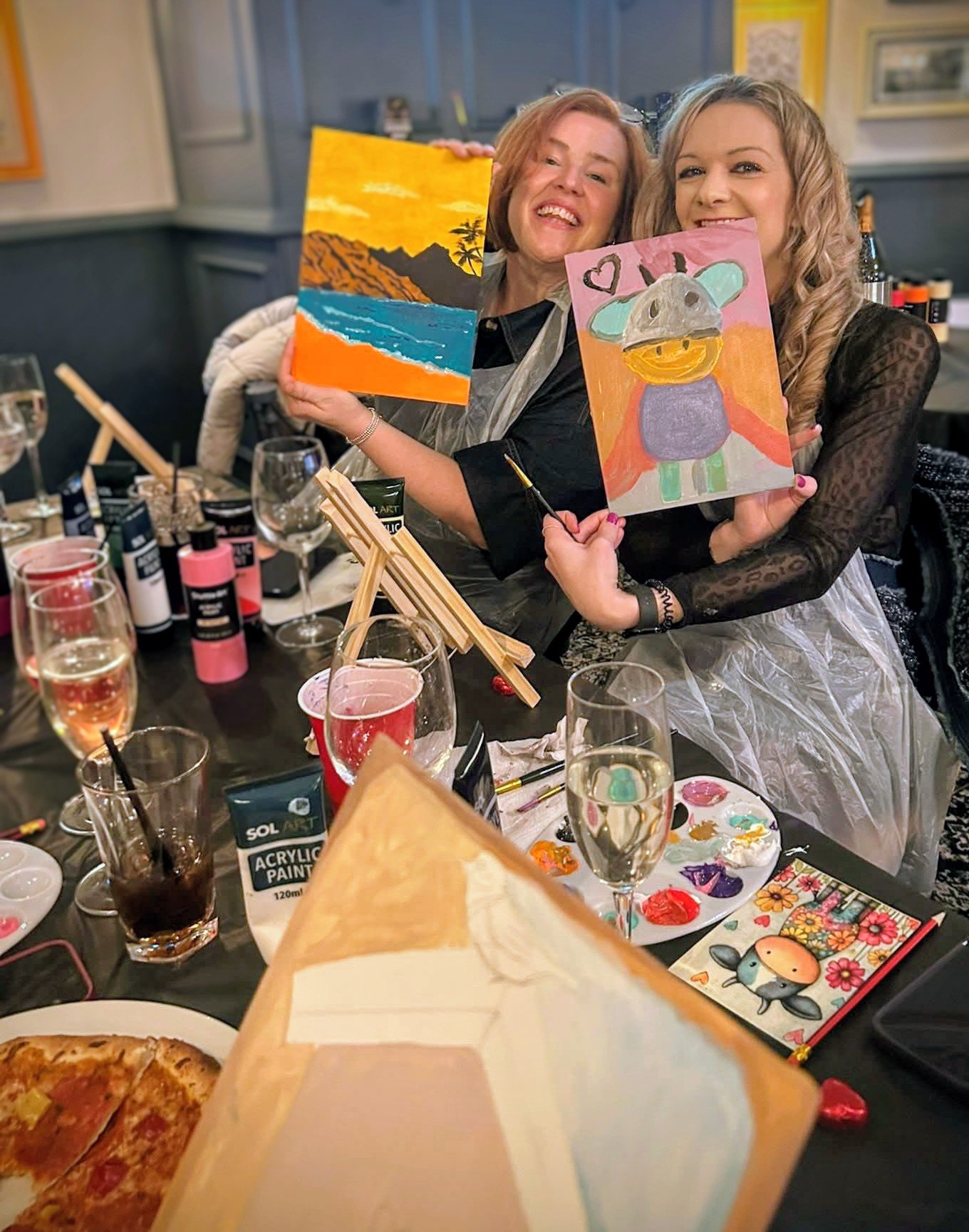 Two women smiling at a paint and sip class, holding up their colourful artworks, with a table of drinks, a pizza, and painting supplies in the foreground.