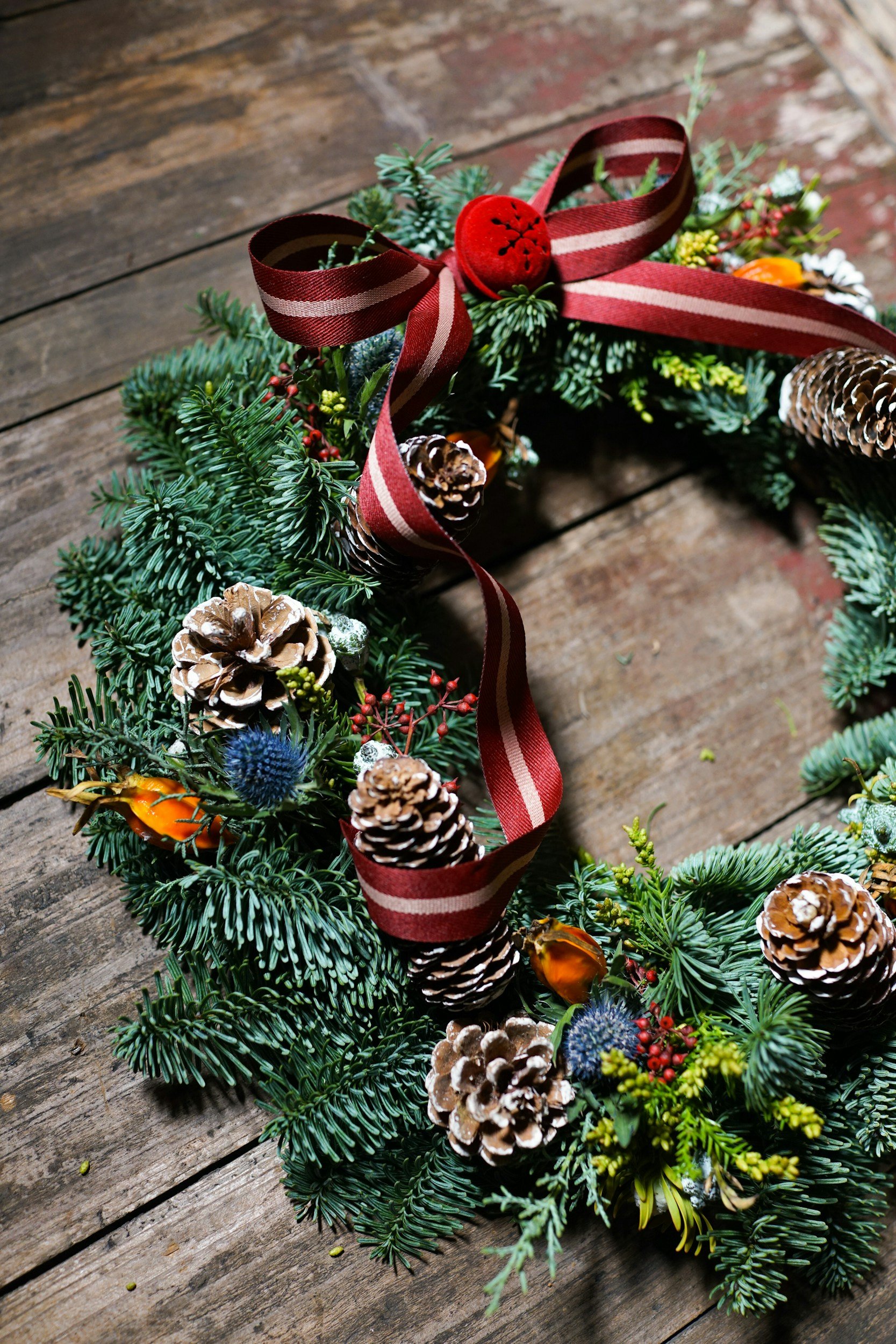 Christmas wreath made of evergreen branches decorated with pinecones, a red and beige ribbon, berries, and dried orange slices, placed on a wooden surface.