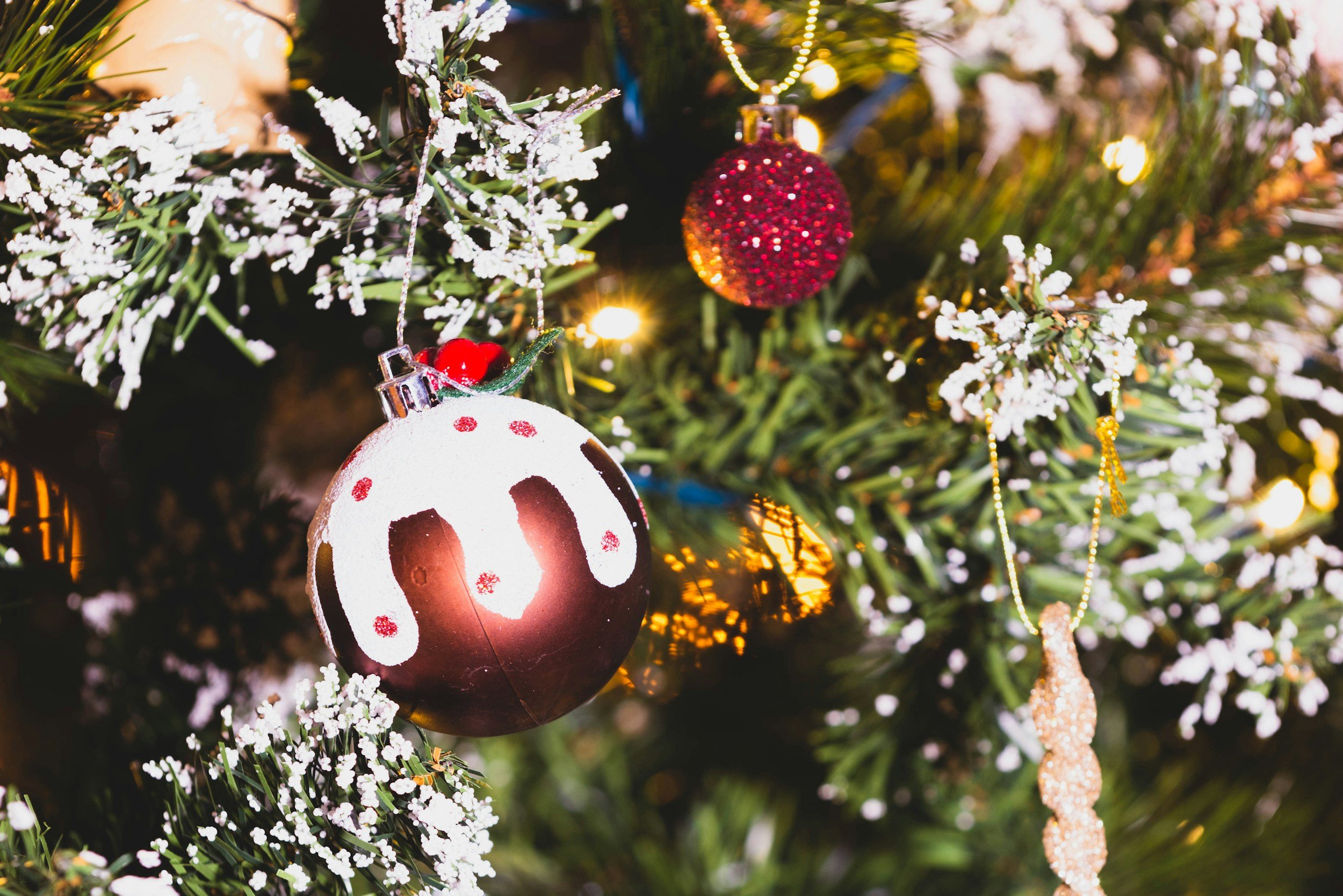 Close-up of a decorated Christmas tree with ornaments, including a round ornament resembling a chocolate ball with white drip design, a red glittery ball, and a golden glittery ornament, among artificial snow, string lights, and green pine branches.