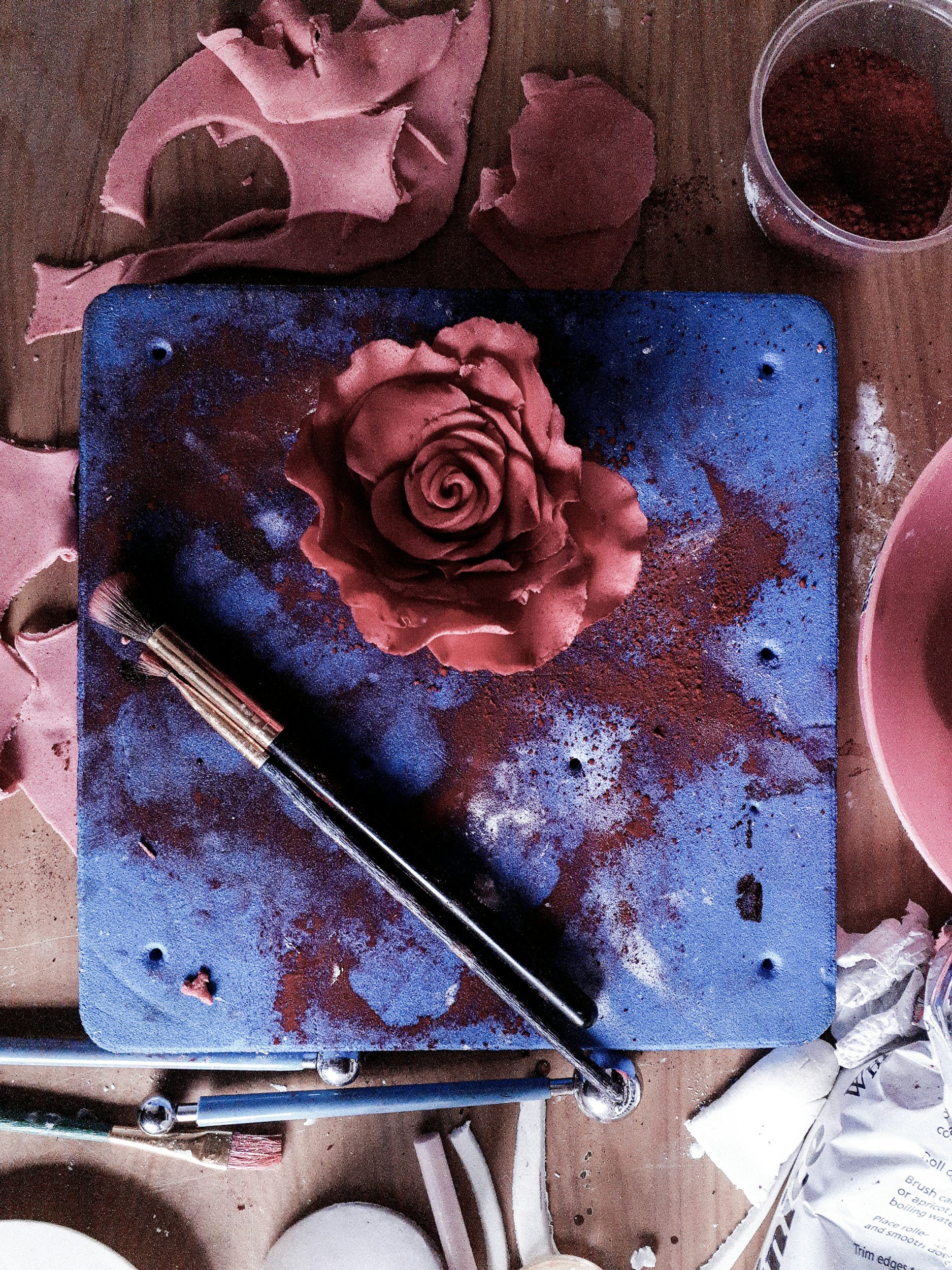 A woodworking workspace with a blue work surface, a rose-shaped sculpture made from pink material, a paintbrush, and scattered pink and brown material, with a container of pink powder or pigment nearby.