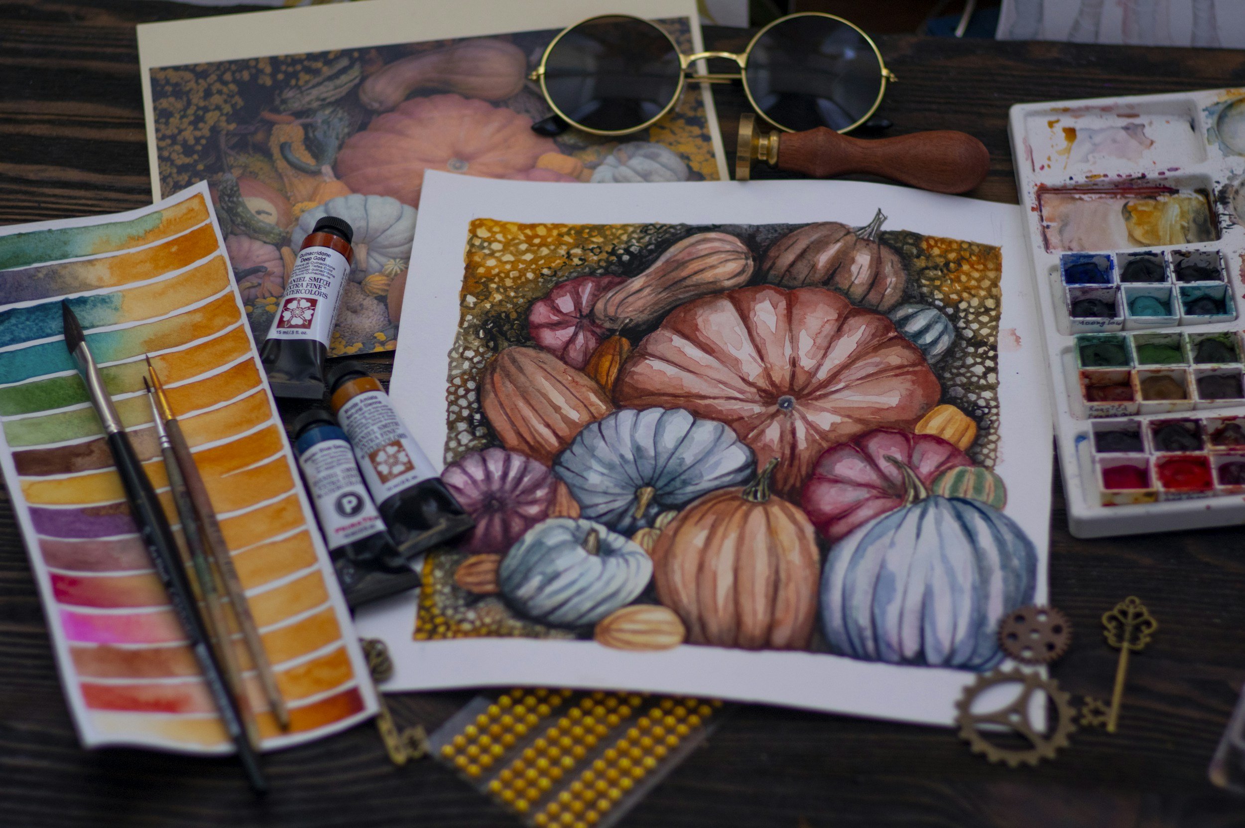 A watercolor painting of various pumpkins and gourds on paper, surrounded by watercolor paints, brushes, color swatches, and art tools on a wooden table.