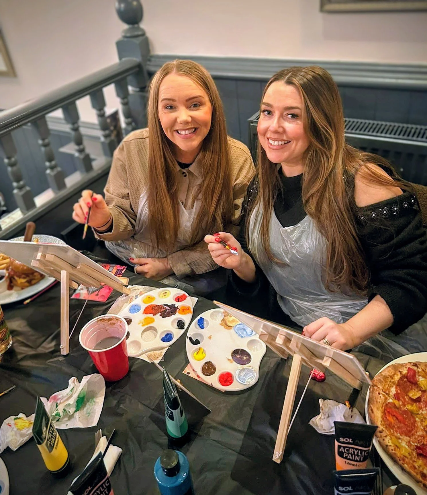 Two women are at a table painting at a paint and sip party, wearing protective aprons, surrounded by painting supplies, food, and beverages, smiling at the camera.