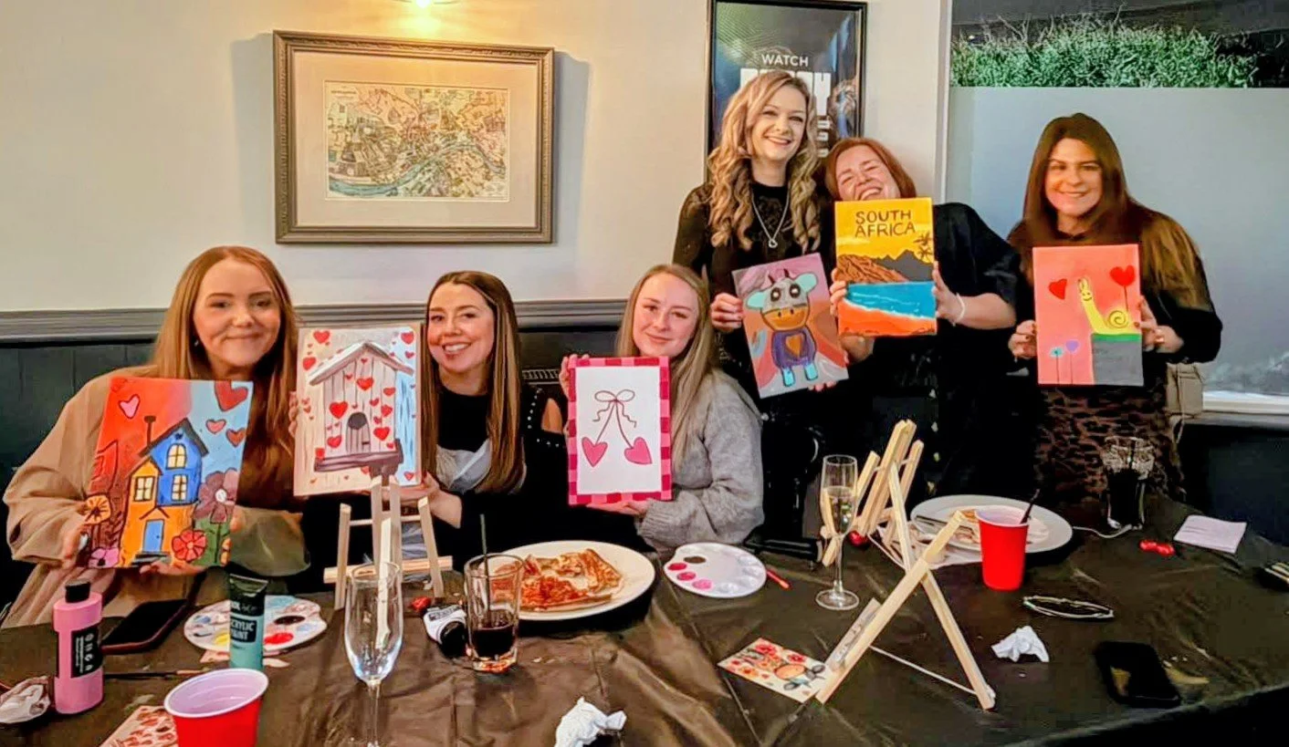 Group of six women displaying painted canvases at a table inside a pub at a paint and sip party, with painting supplies and a pizza, some holding colourful artworks of animals, landscapes, and abstract designs.