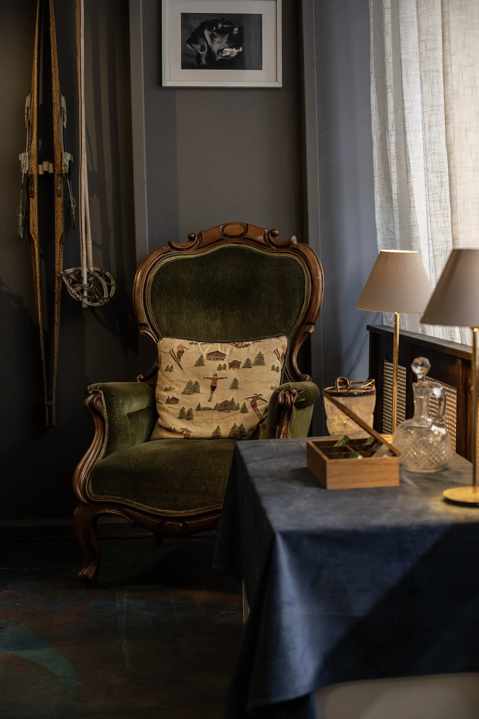 A vintage green upholstered armchair with wooden frame, a patterned cushion featuring nature scenes, against a dark wall. Above, a black-and-white framed photograph of a dog. To the right, a window with sheer curtains, a table with lamps, and decorative glass containers.
