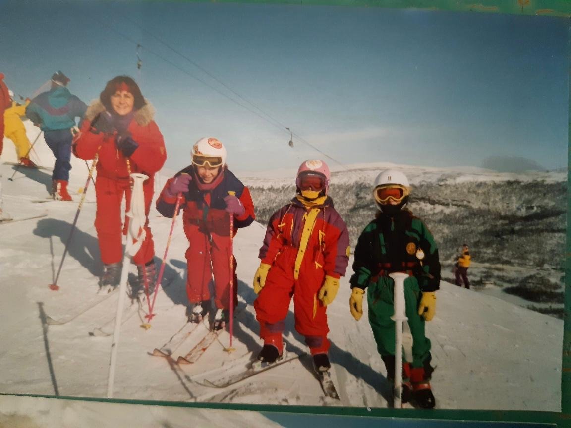A group of children posing in ski gear on a snowy mountain with ski slopes and a ski lift cable in the background.