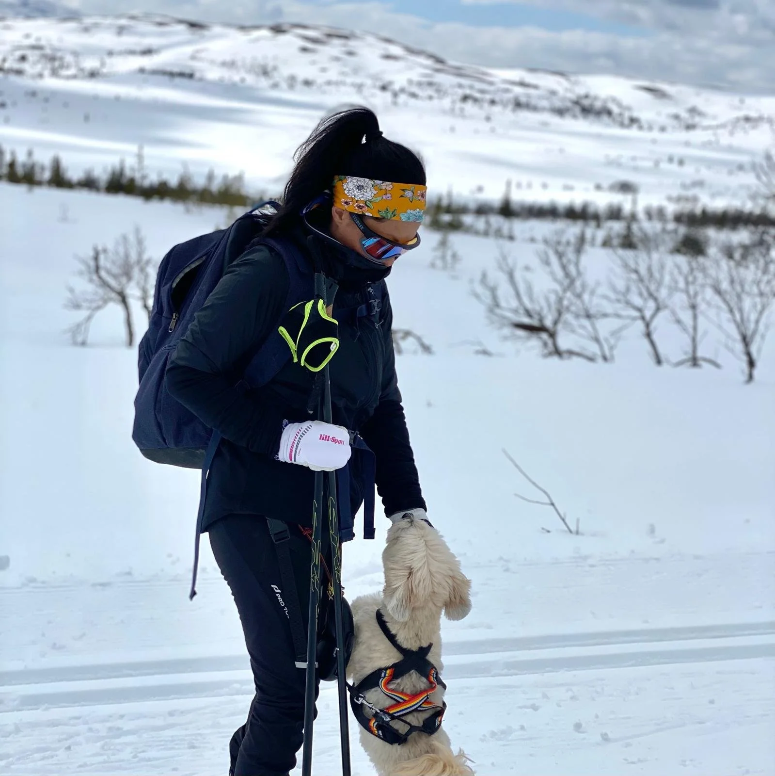 Person with long dark hair, wearing a colorful eye mask and yellow headband, standing in the snow with a guide dog, holding a walking stick, and wearing dark winter gear, in a snowy landscape with some sparse trees and hills in the background.