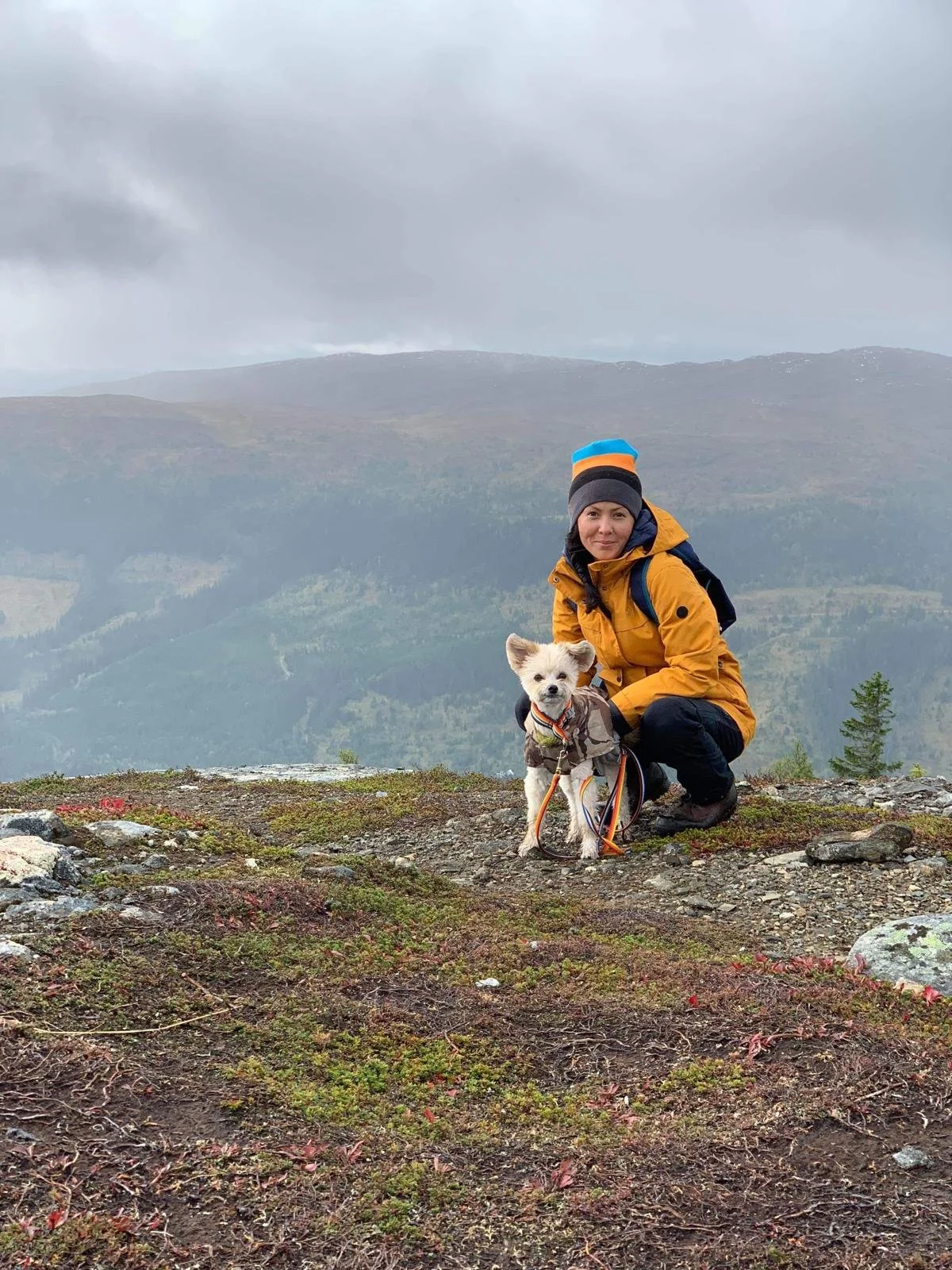 A woman hiking on a mountain trail with her small dog, both wearing outdoor gear, with a scenic mountain landscape and cloudy sky in the background.