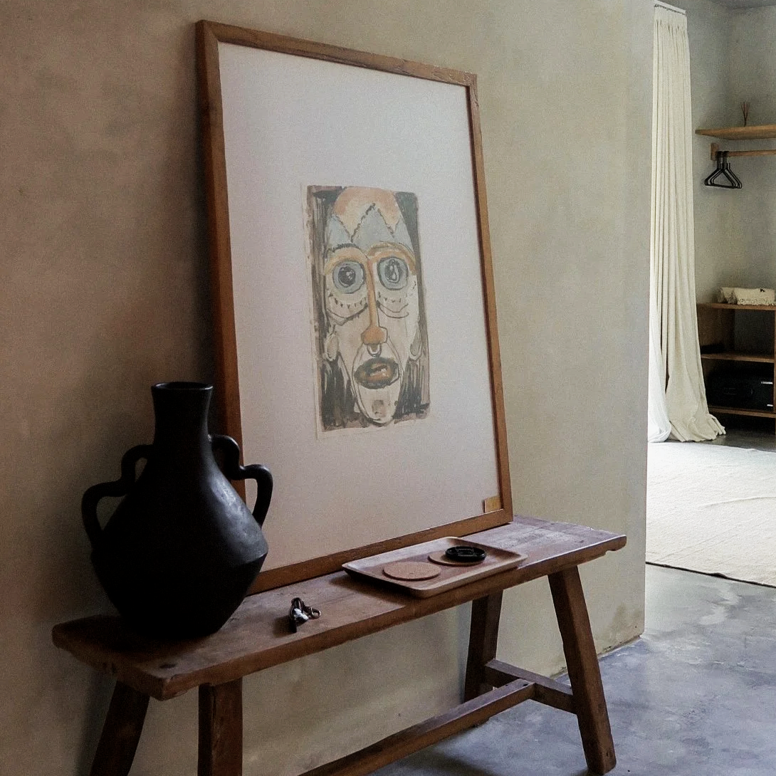A framed abstract artwork on an easel, with a black ceramic vase, keys, and coasters on a wooden table beside it in a living space.