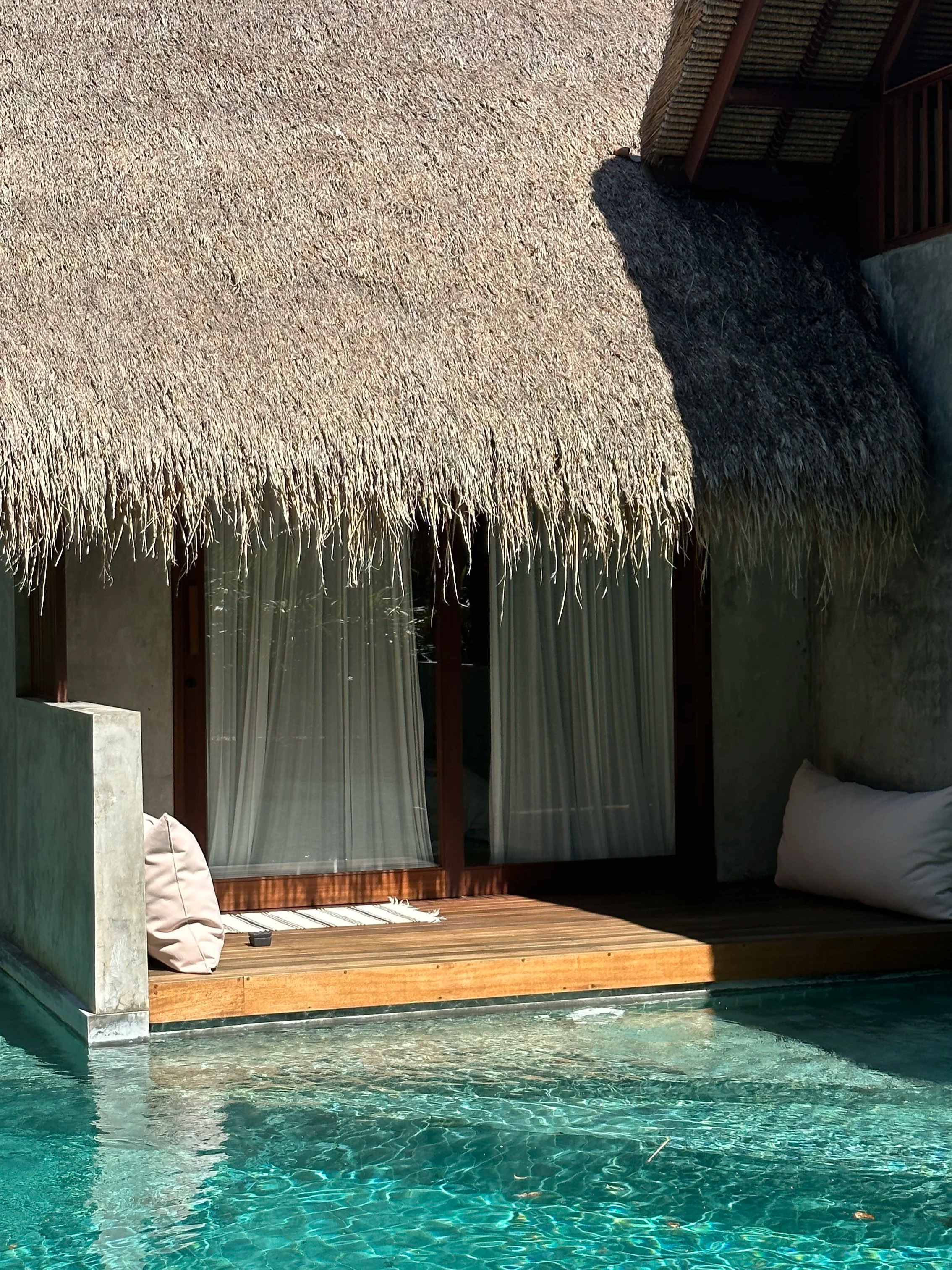 Overwater bungalow with thatched roof, glass doors with curtains, wooden deck with cushions, beside a swimming pool.