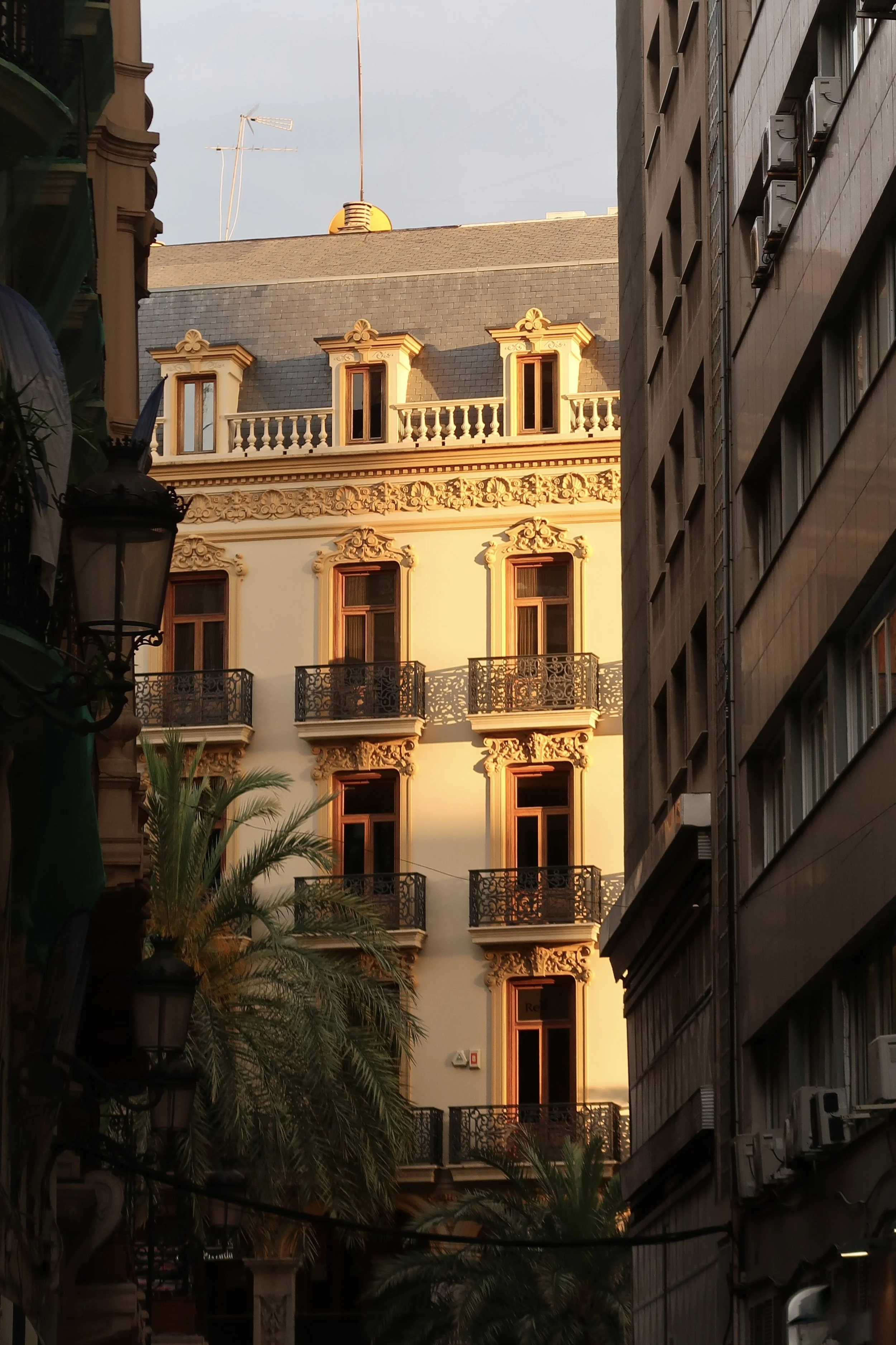 A historic building with ornate architectural details and balconies, illuminated by the warm glow of sunlight, surrounded by modern buildings and palm trees.