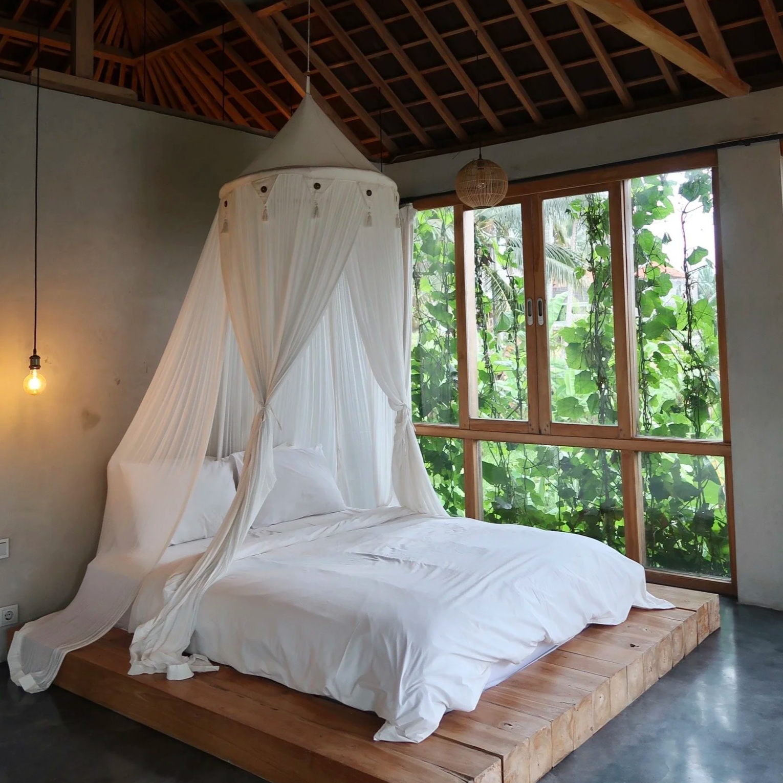 A cozy bedroom with a four-poster bed draped with white fabric, a wooden platform base, large window with green foliage outside, a hanging light bulb, and woven ceiling accents.