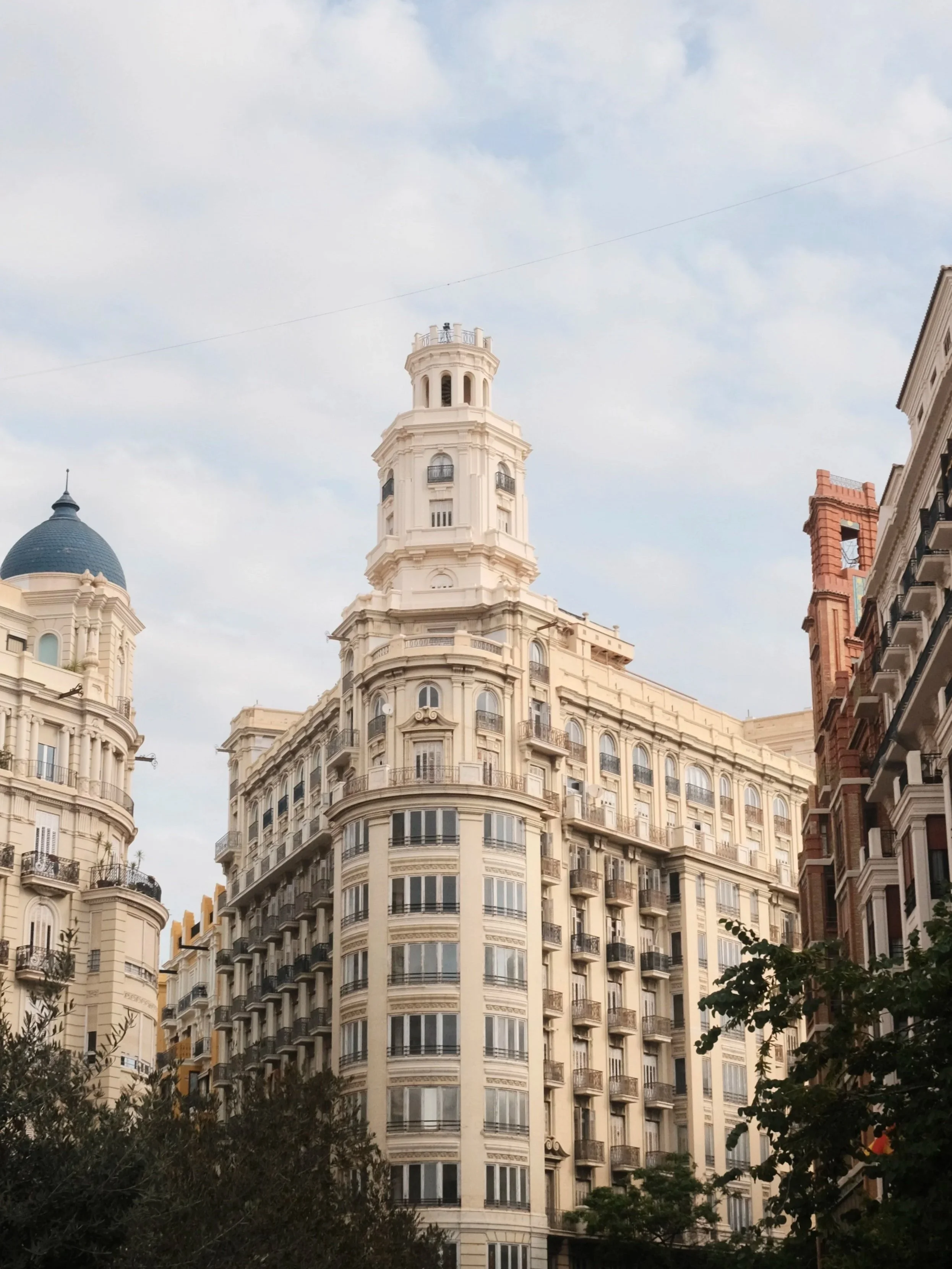 Tall historical building with a decorative white facade, multiple windows, and a tower at the top, set against a partly cloudy sky. Valencia, Spain.