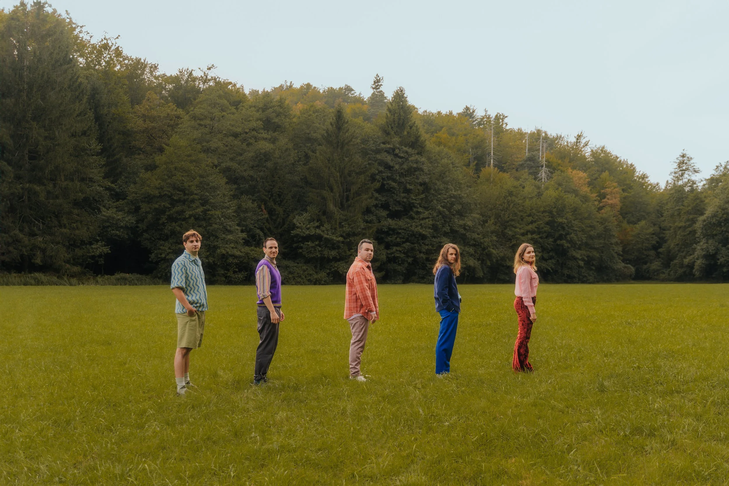Five people standing in a line on a grassy field with a forest in the background.
