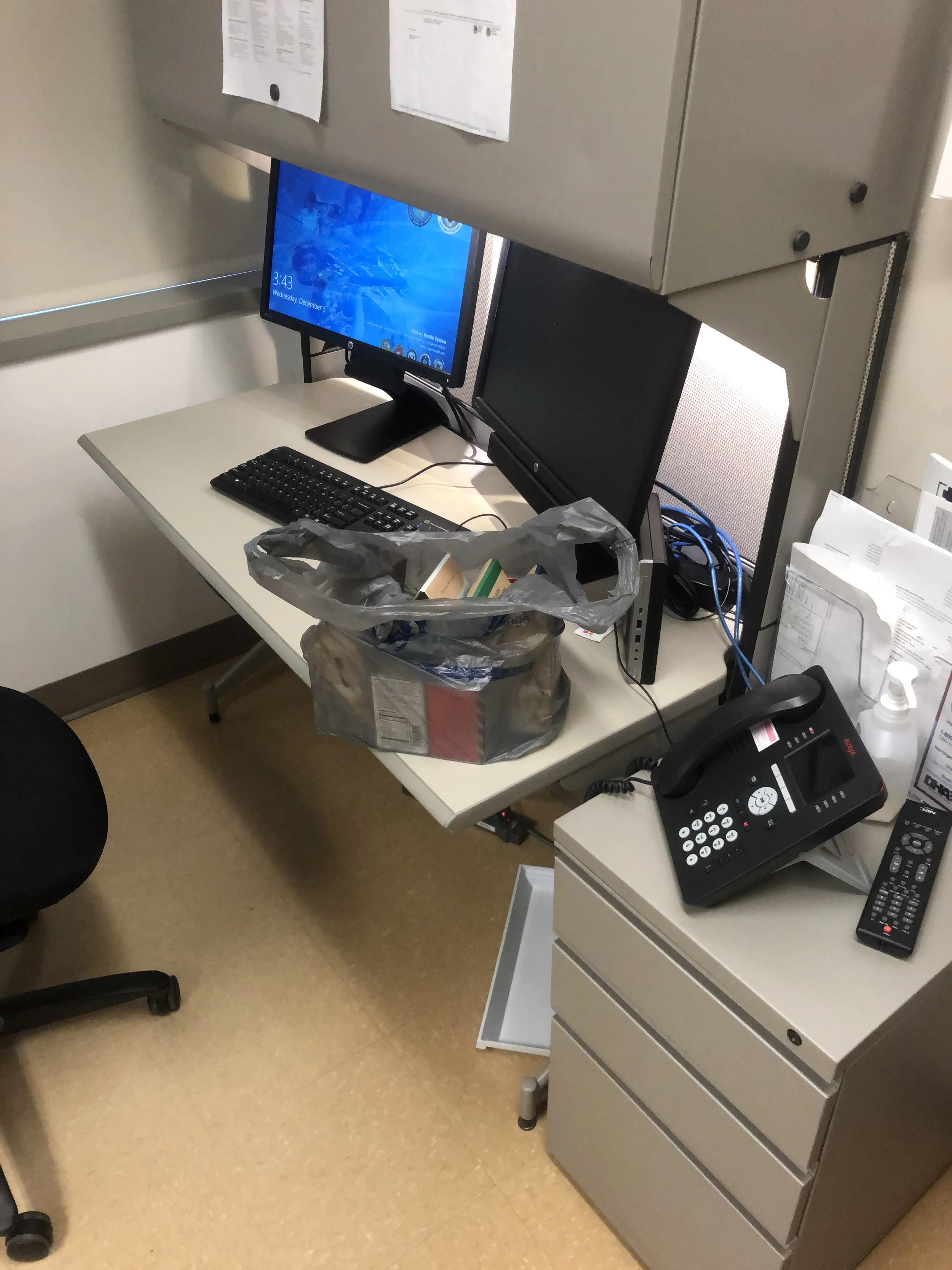 Office cubicle with a desk, computer monitor, keyboard, and a plastic bag filled with items on the desk. There is a legal phone on a side table, and papers tacked on a metal cabinet. The floor is beige, and the workspace appears cluttered.